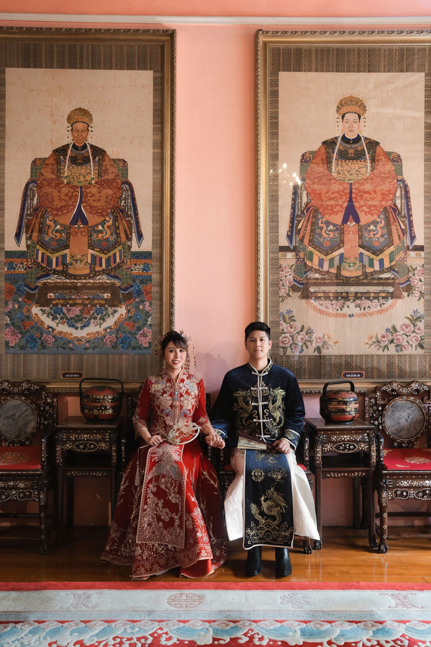 Wedding couple in traditional Chinese attire sitting below ancestral paintings at the Pinang Peranakan Mansion