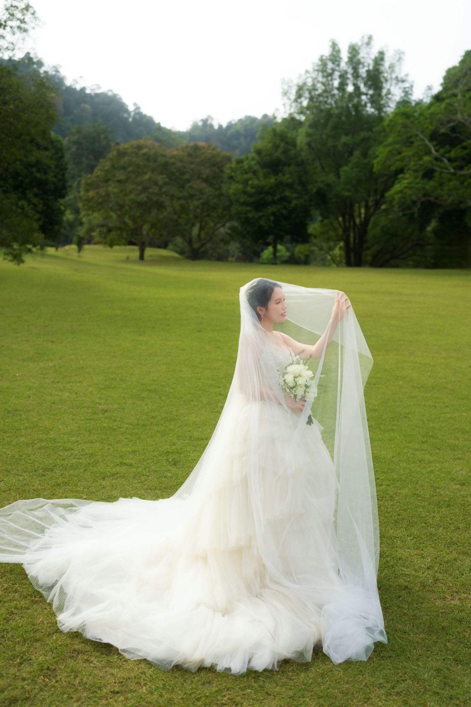 Bride in flowing white gown poses gracefully on a green lawn with her veil flowing in the wind.