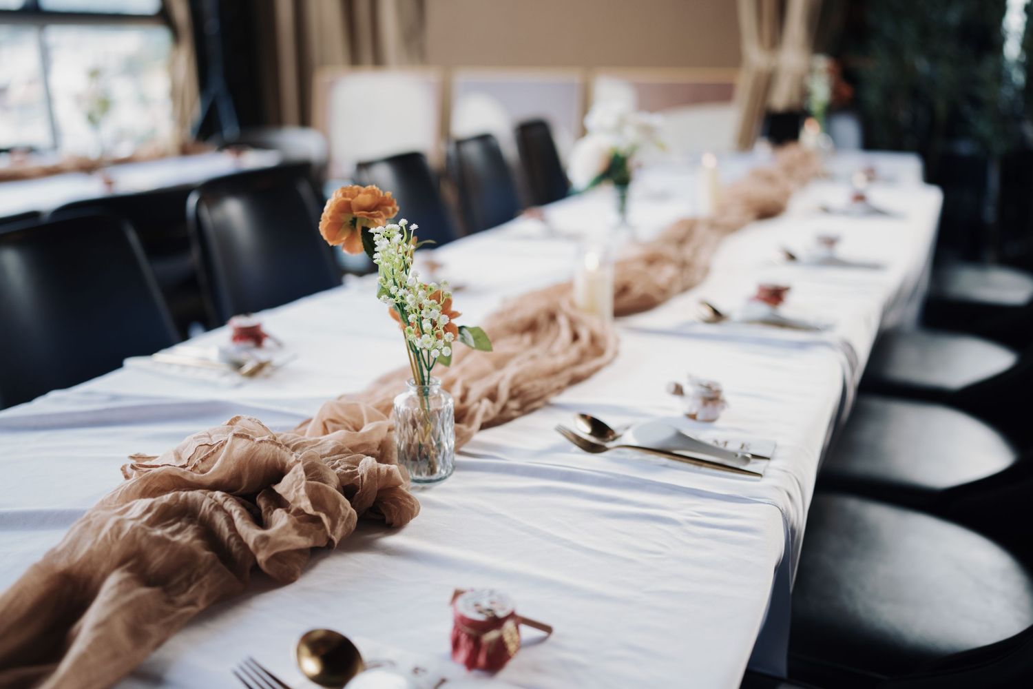 Elegant boho wedding table decor featuring a tan cheesecloth runner, gold cutlery, and rustic floral centerpieces.