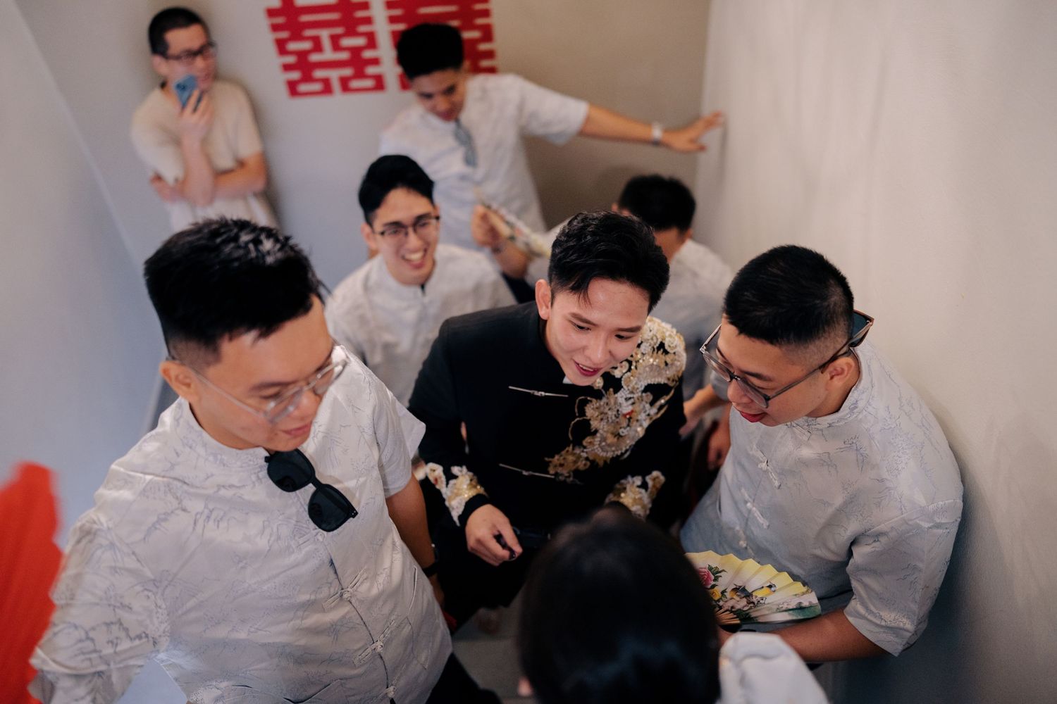 A groom in a black embroidered jacket surrounded by groomsmen in blue shirts on a staircase
