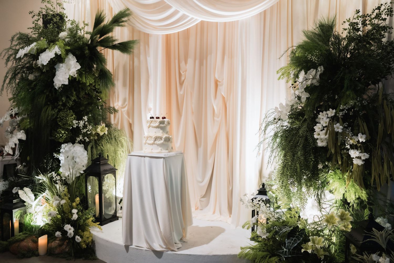 Elegant wedding backdrop with white drapes and lush greenery floral arrangements.