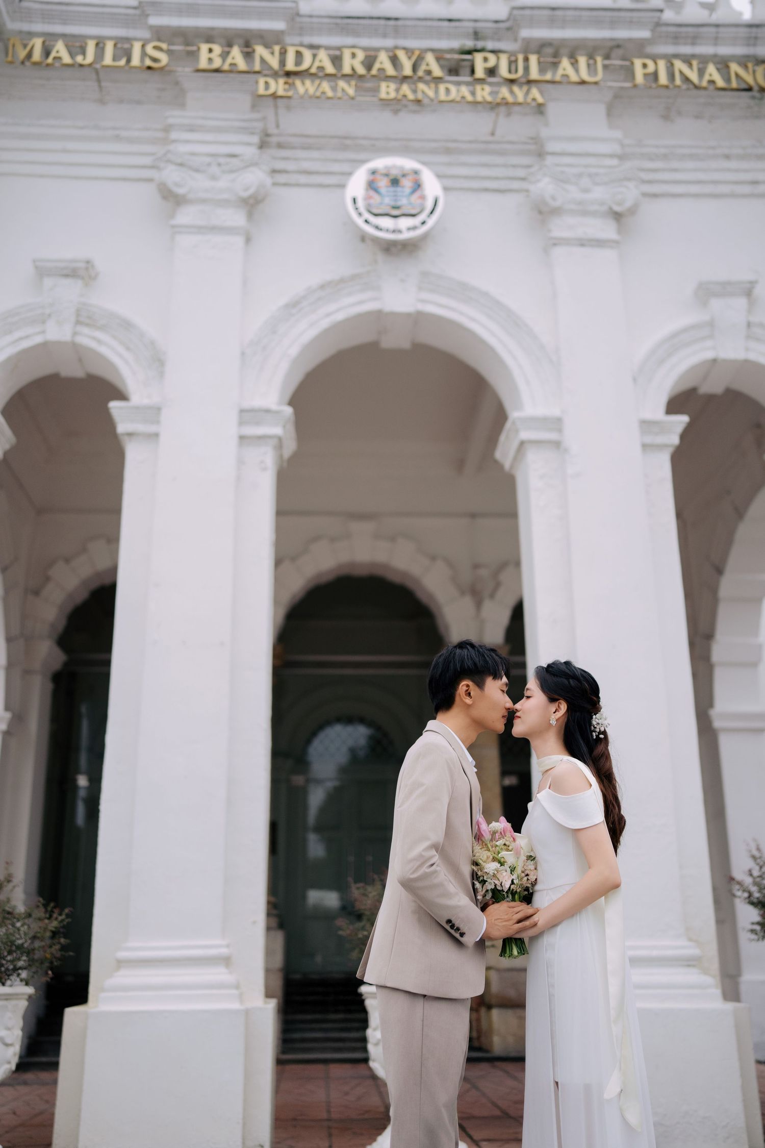A romantic wedding photo in front of the Majlis Bandaraya Pulau Pinang building with white colonial architecture.