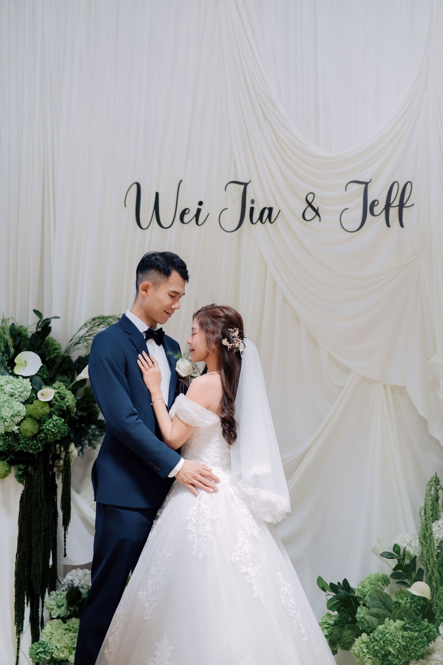 Couple embracing before a white fabric backdrop with black cursive names and floor florals