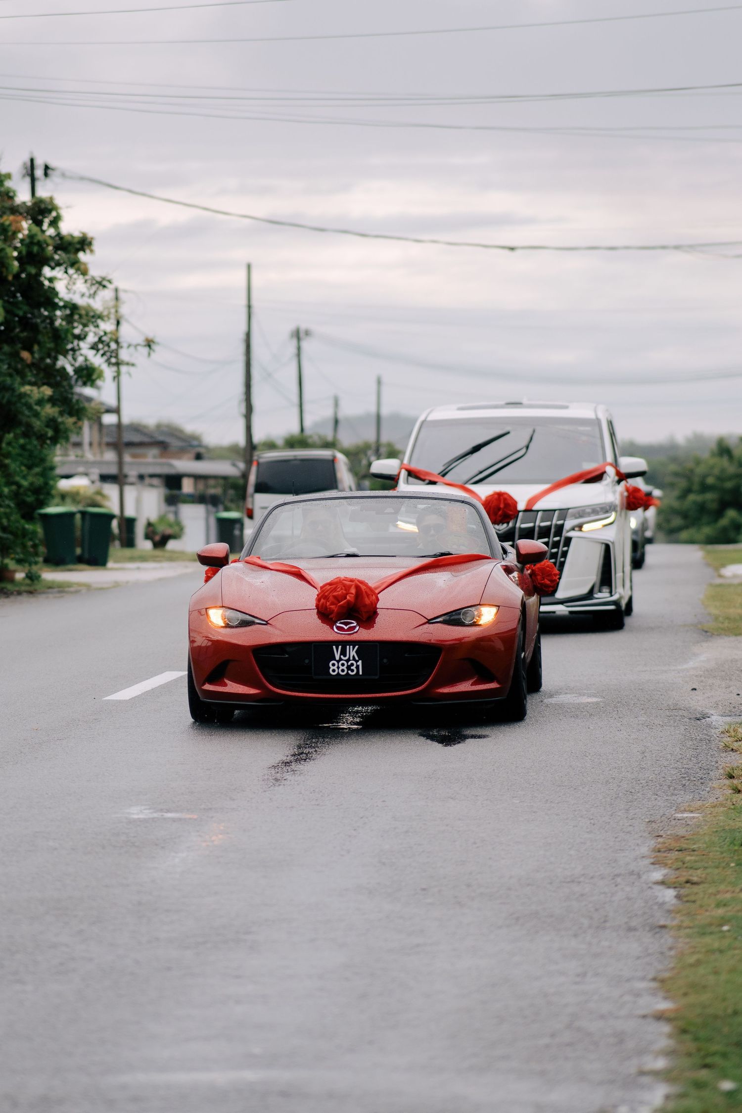 A red convertible wedding car decorated with a large red ribbon leading a procession on a Penang street