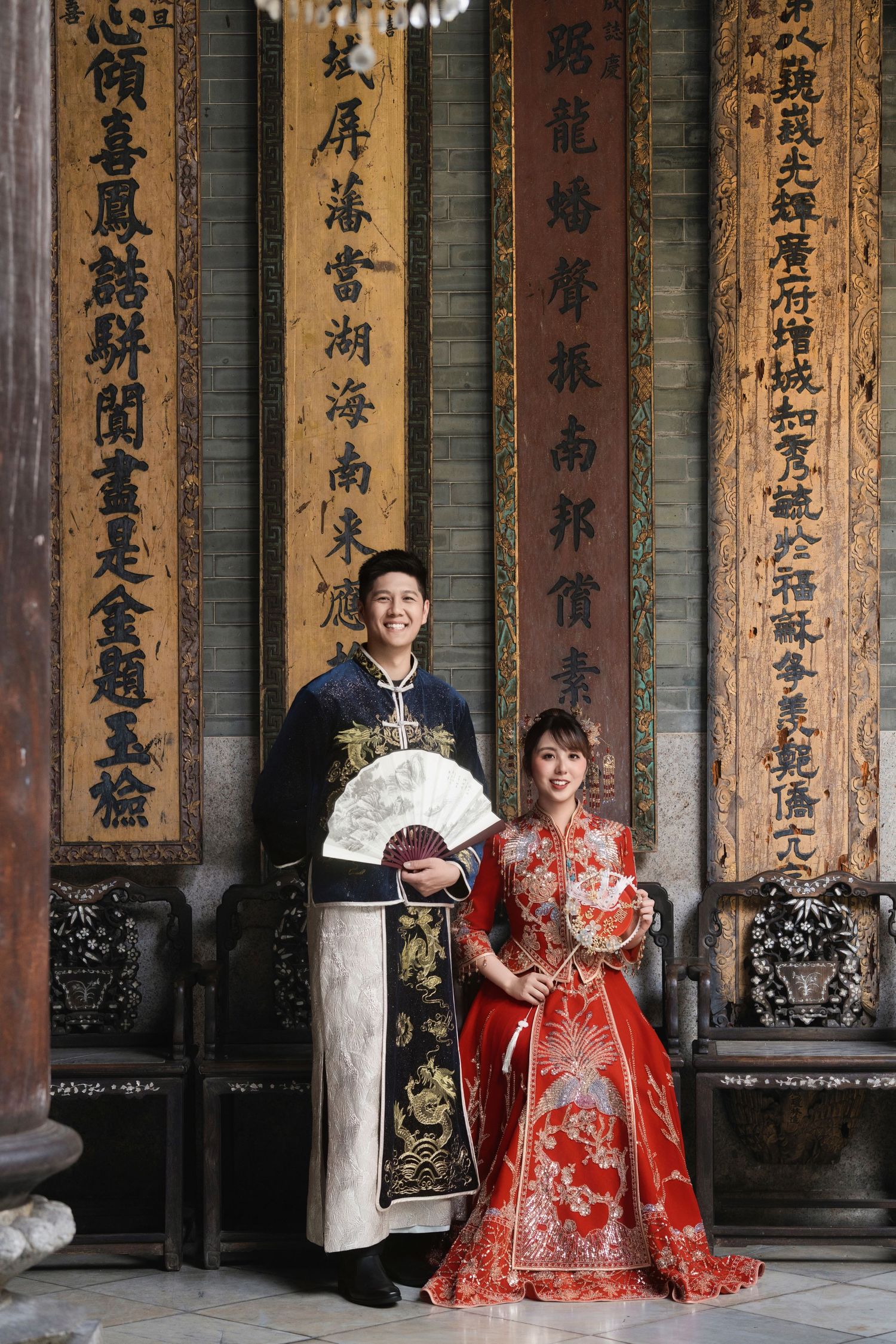 Groom standing with a white fan next to the bride sitting in a red gown, both in traditional Chinese wedding attire