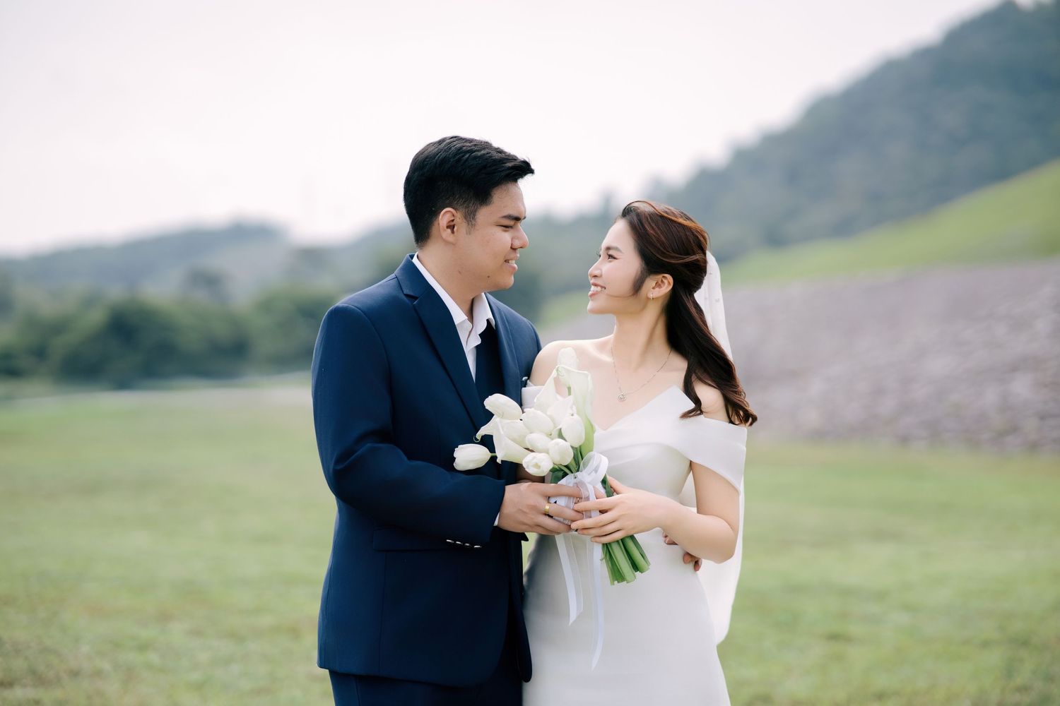 Bride and groom in wedding attire looking at each other, holding a white tulip bouquet at Mengkuang Dam