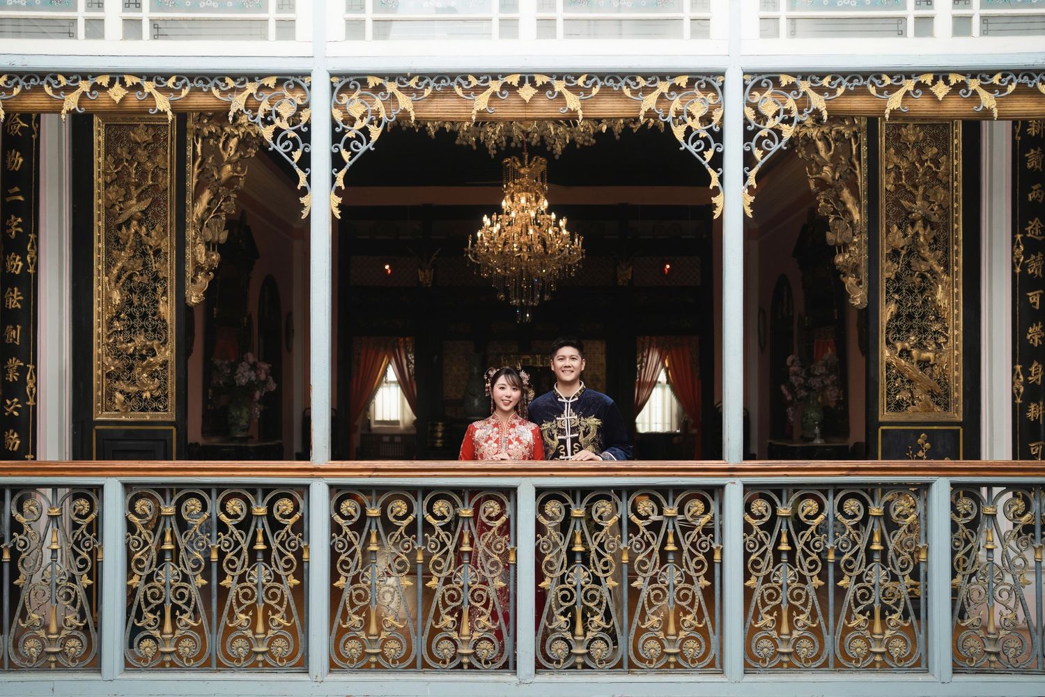 Couple in traditional wedding dress standing on a balcony with intricate gold carvings and Peranakan architectural details