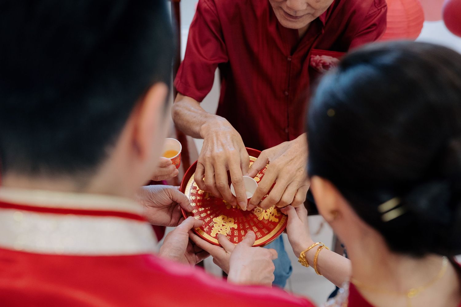 Couple practicing Chinese tea ceremony traditions by serving tea on a red and gold wedding tray to a seated elder