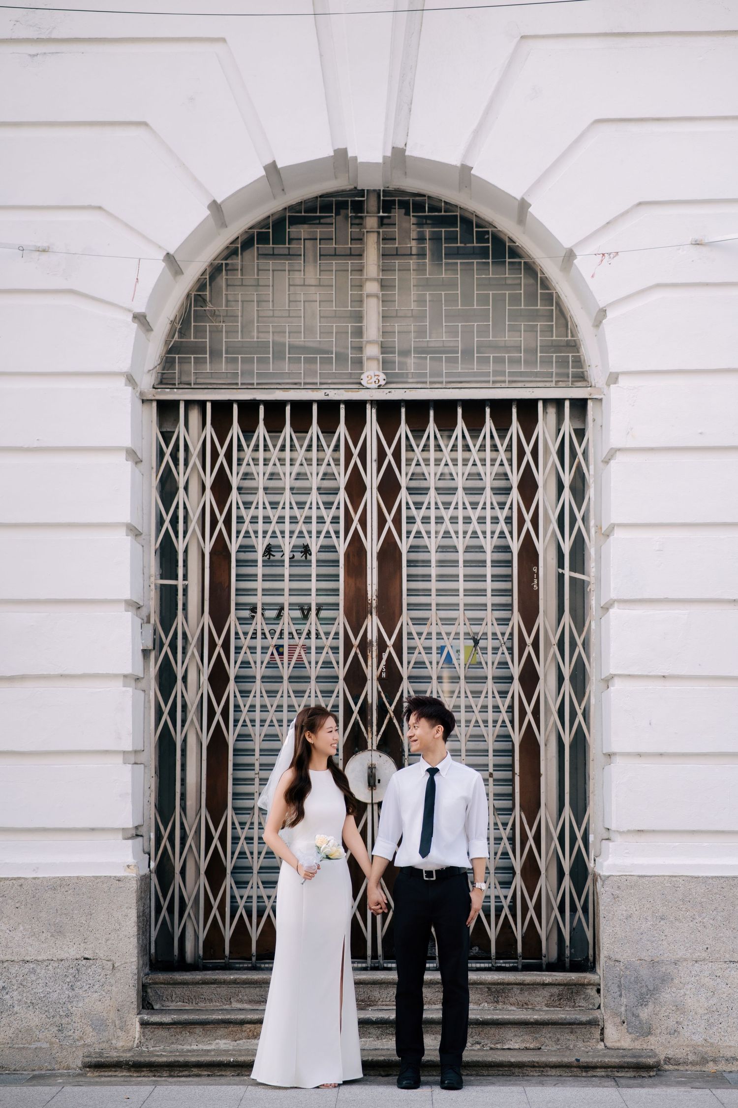 A couple poses playfully in front of an ornate white archway with decorative metal grating.
