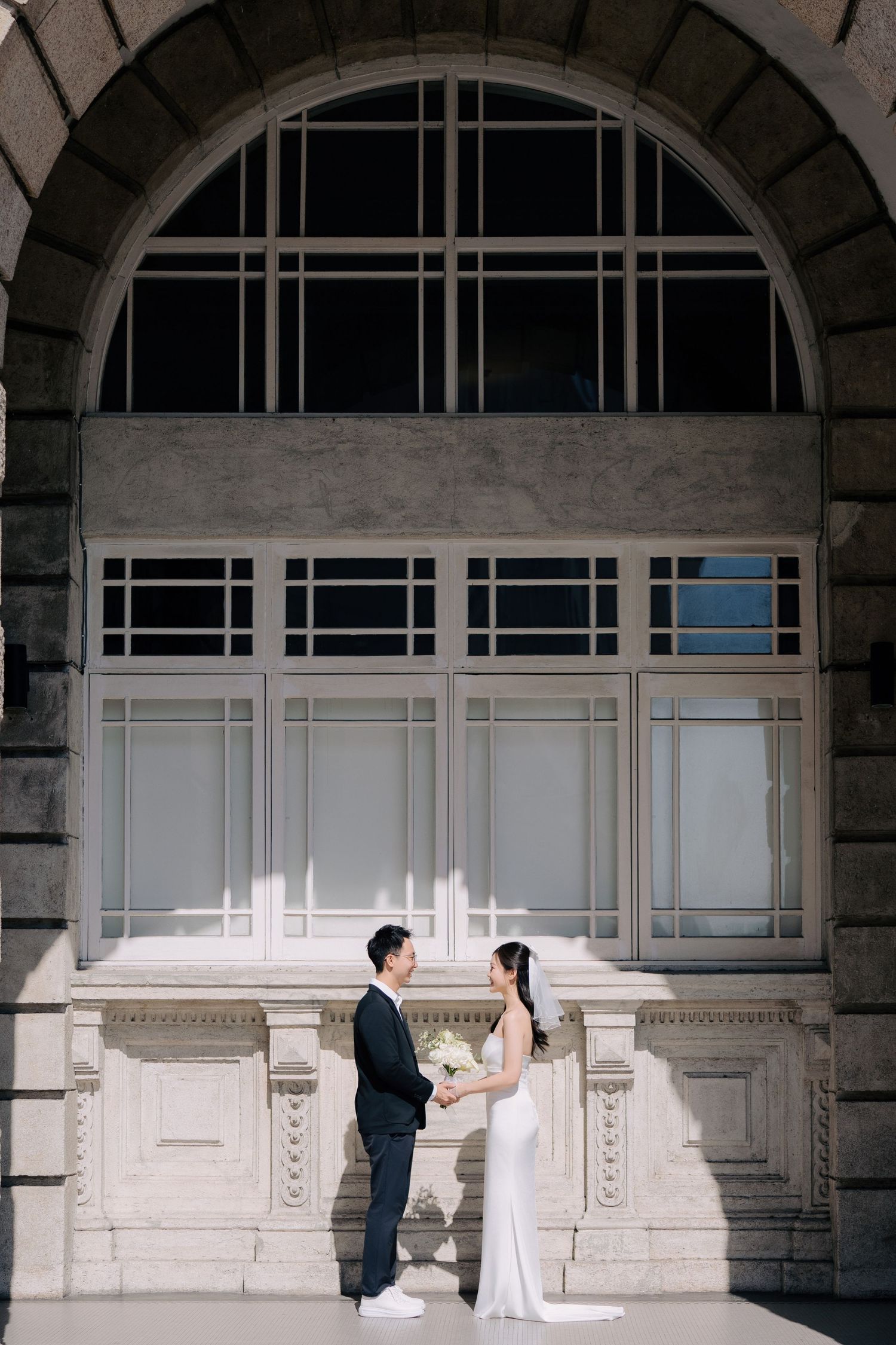 A couple poses against an architectural backdrop with arched windows and white stone details.