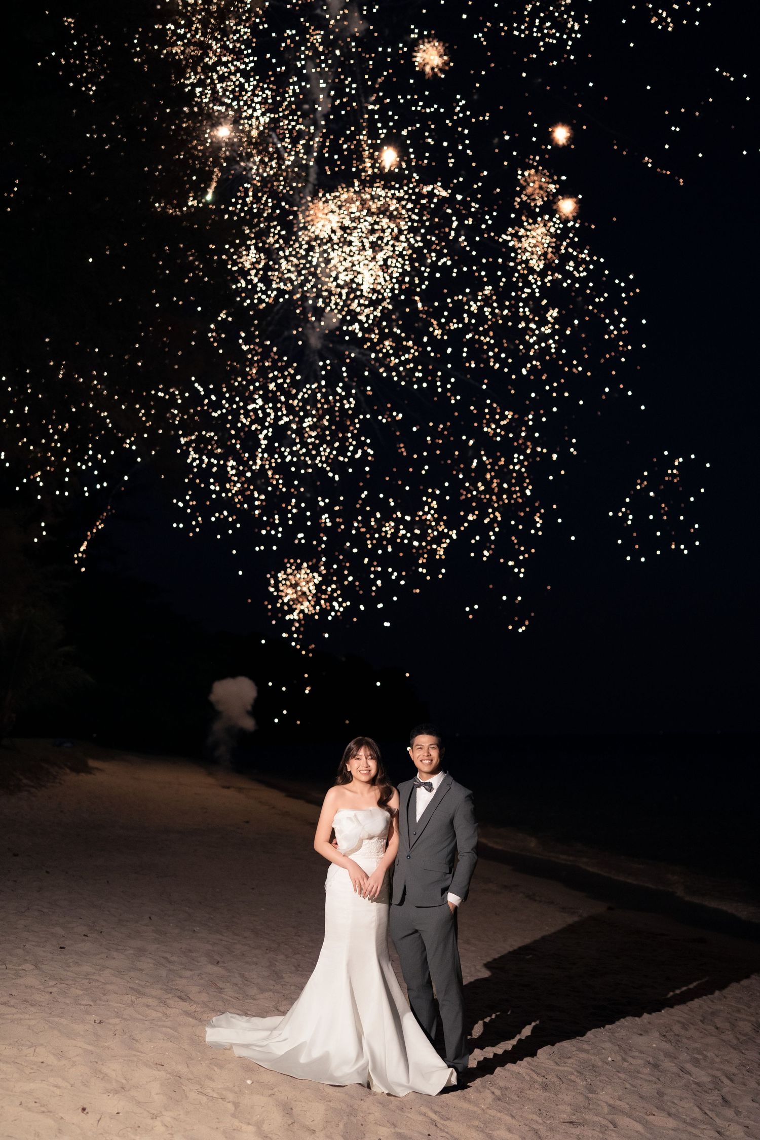 Wedding couple silhouetted against spectacular fireworks display on a sandy beach at night.