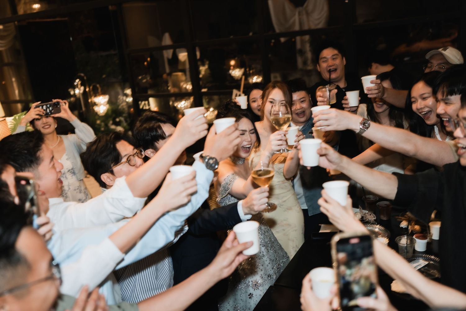 Wedding after-party celebration featuring a bride and guests raising cups for a casual reception toast (yam seng) at an indoor night venue
