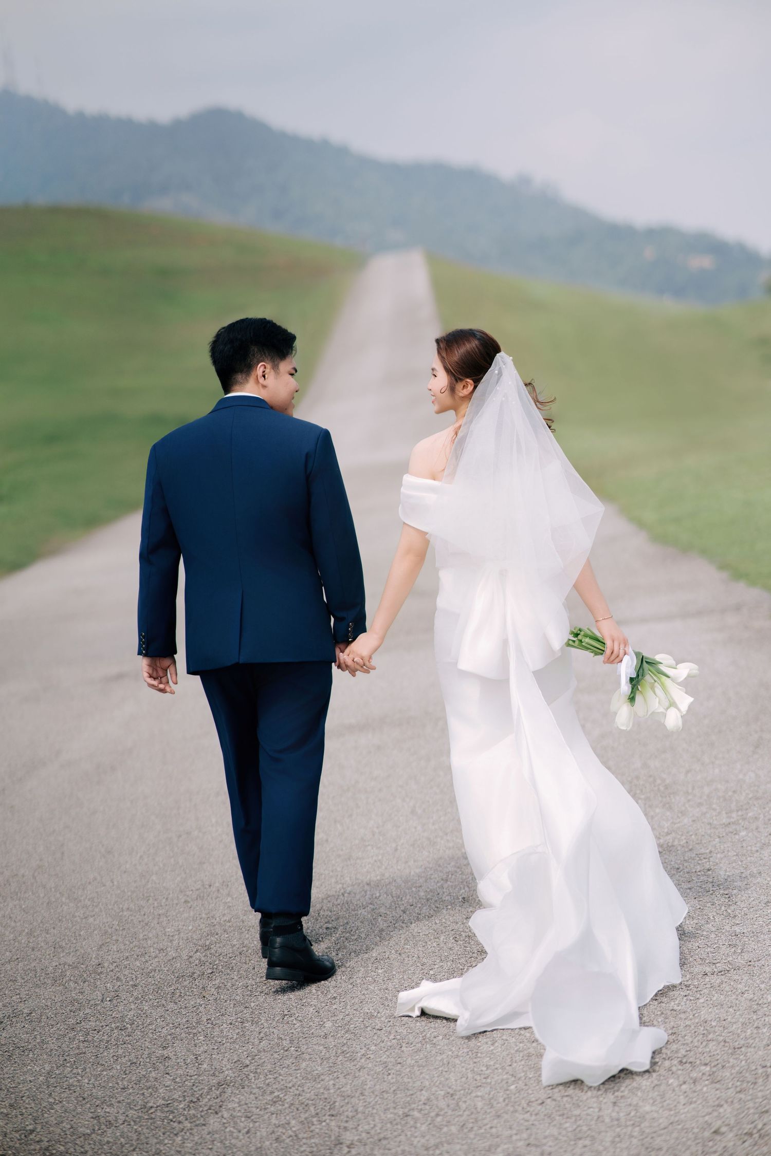 A couple walks down a winding road with flowing wedding dress and veil in the breeze.