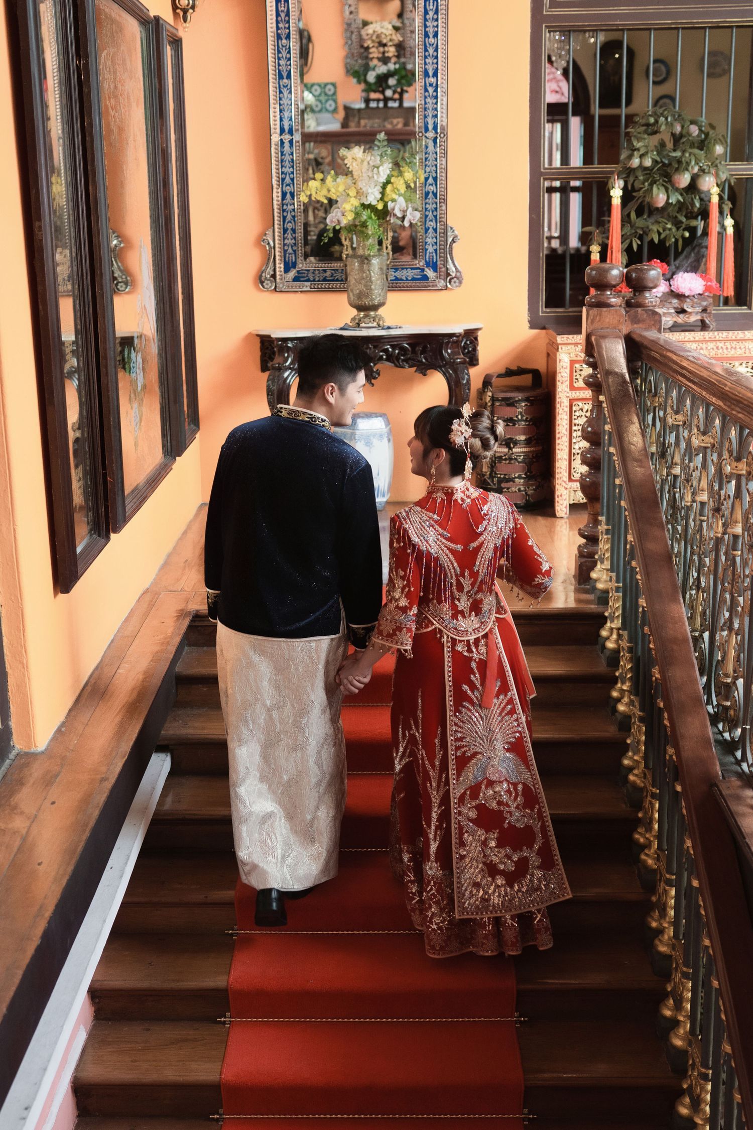 Couple in ornate ceremonial robes walking hand-in-hand up a vintage wooden staircase during their wedding shoot