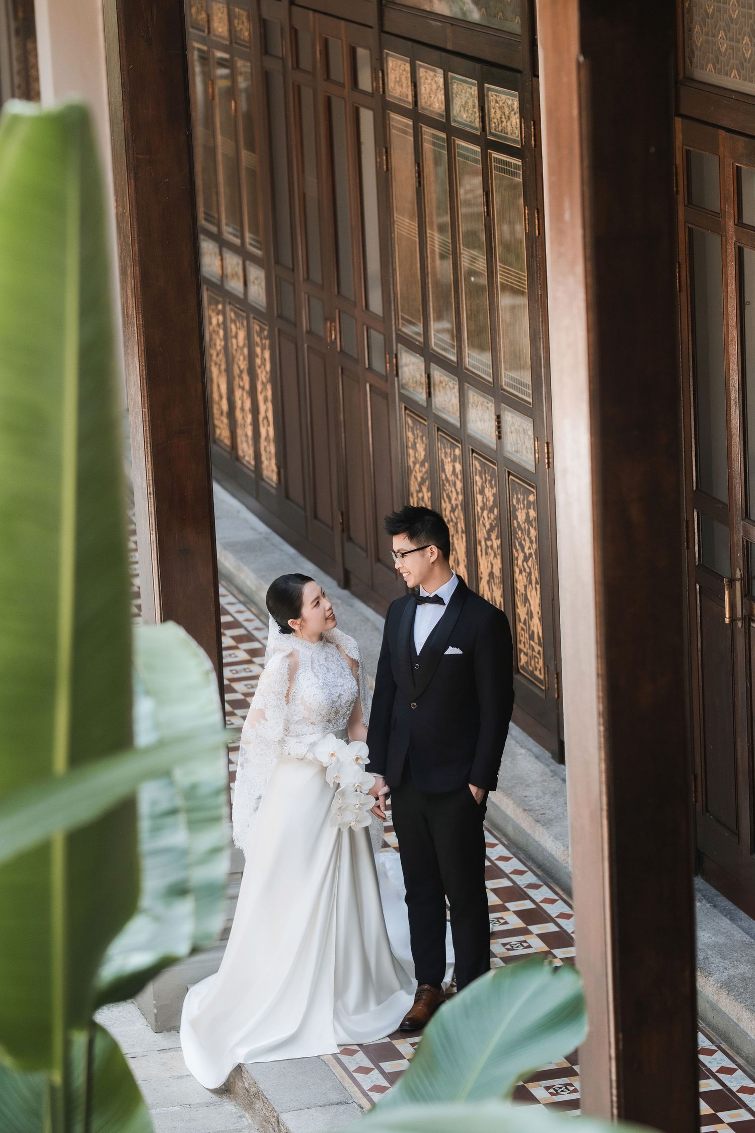 Wedding couple in formal attire posing in an elegant wooden hallway with tropical plants.