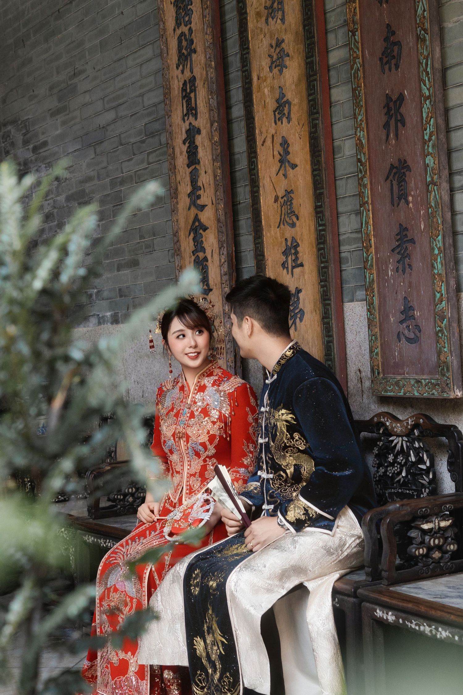 Wedding couple in traditional Chinese attire photographed in front of ornate calligraphy wall.