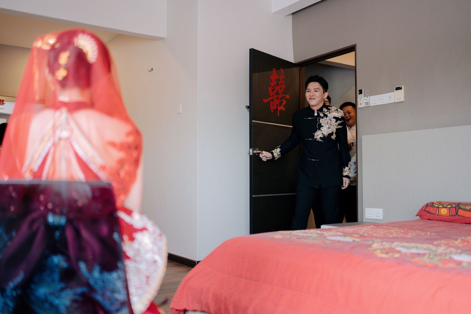Groom in black embroidered cheongsam smiles while opening a door with a red Double Happiness 囍 sticker