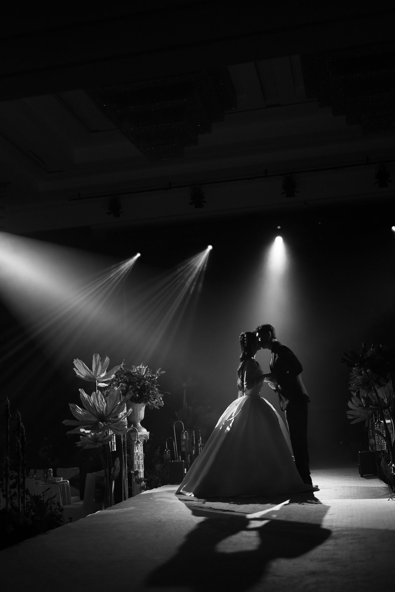 Black and white silhouette of a couple kissing on a wedding aisle under three dramatic spotlights