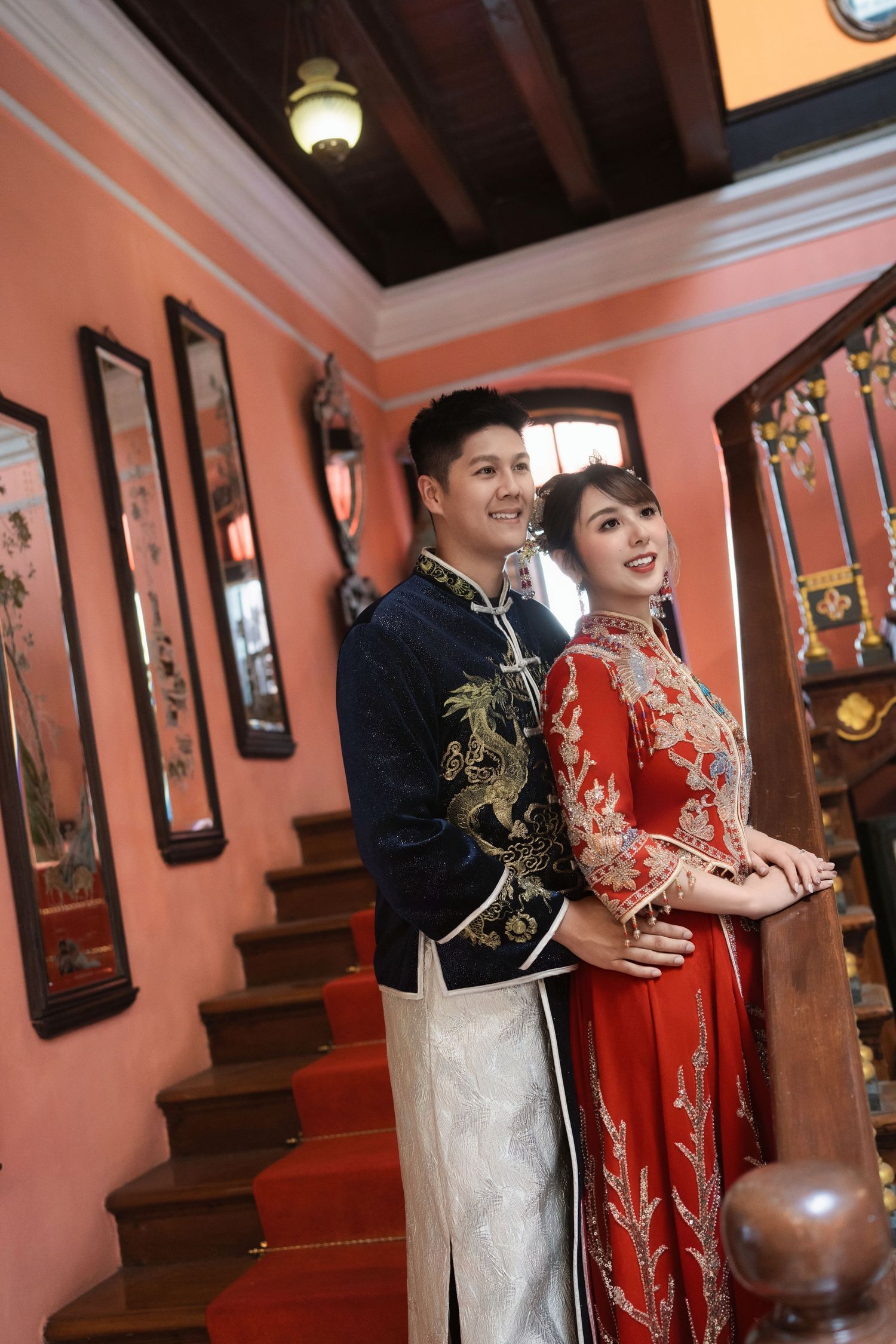 Bride and groom in red and navy ceremonial robes posing on a vintage wooden staircase with red carpeting