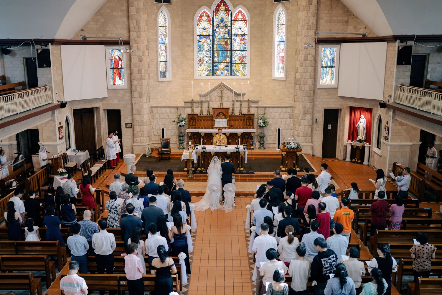 Church wedding ceremony with guests seated in wooden pews under stained glass windows.