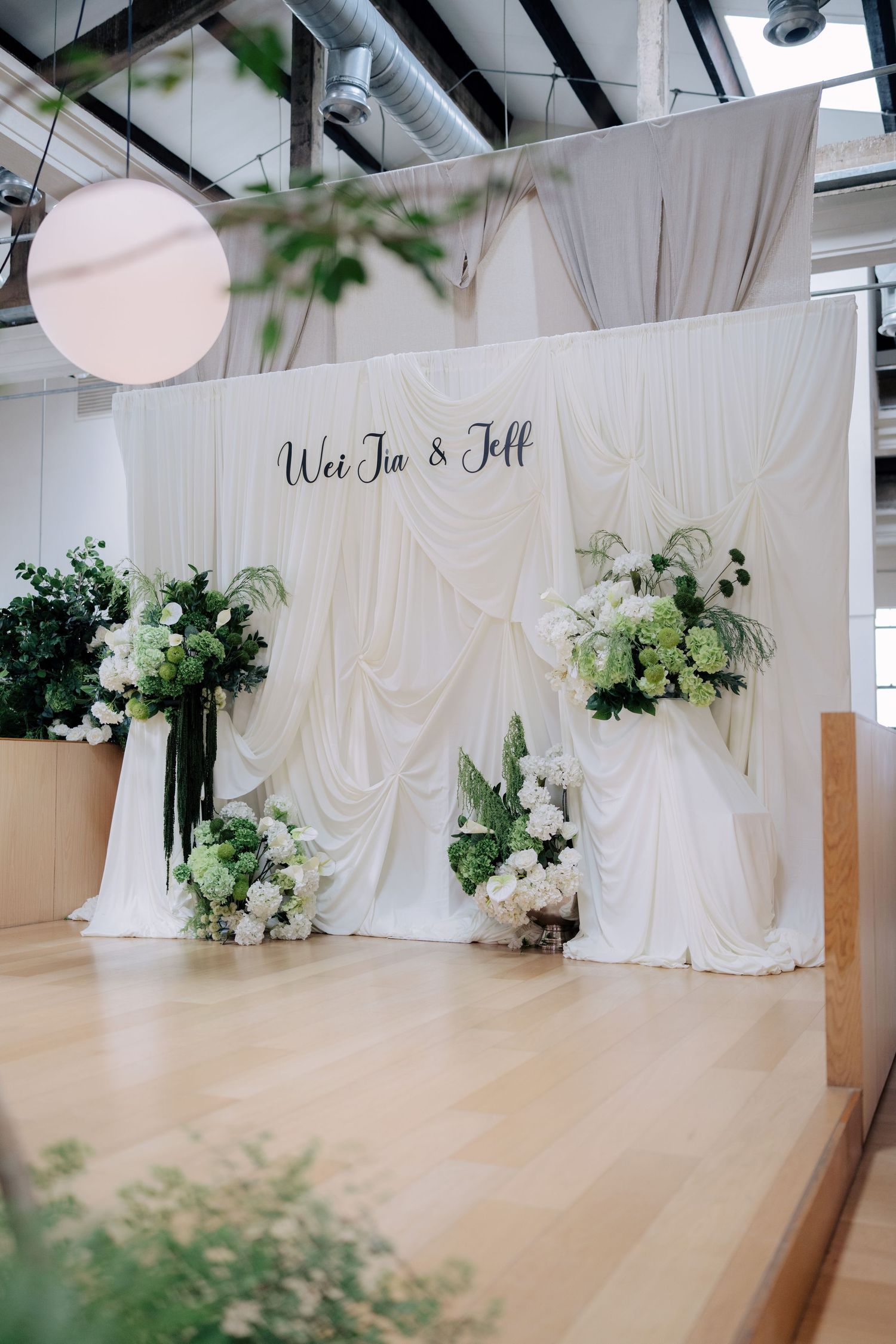 White draped fabric wedding stage backdrop with Wei Jia & Jeff signage and green floral arrangements