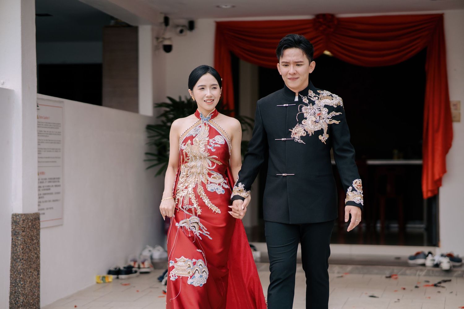 Bride in a backless red dragon-embroidered gown and groom in a matching suit walk hand-in-hand past a red curtain