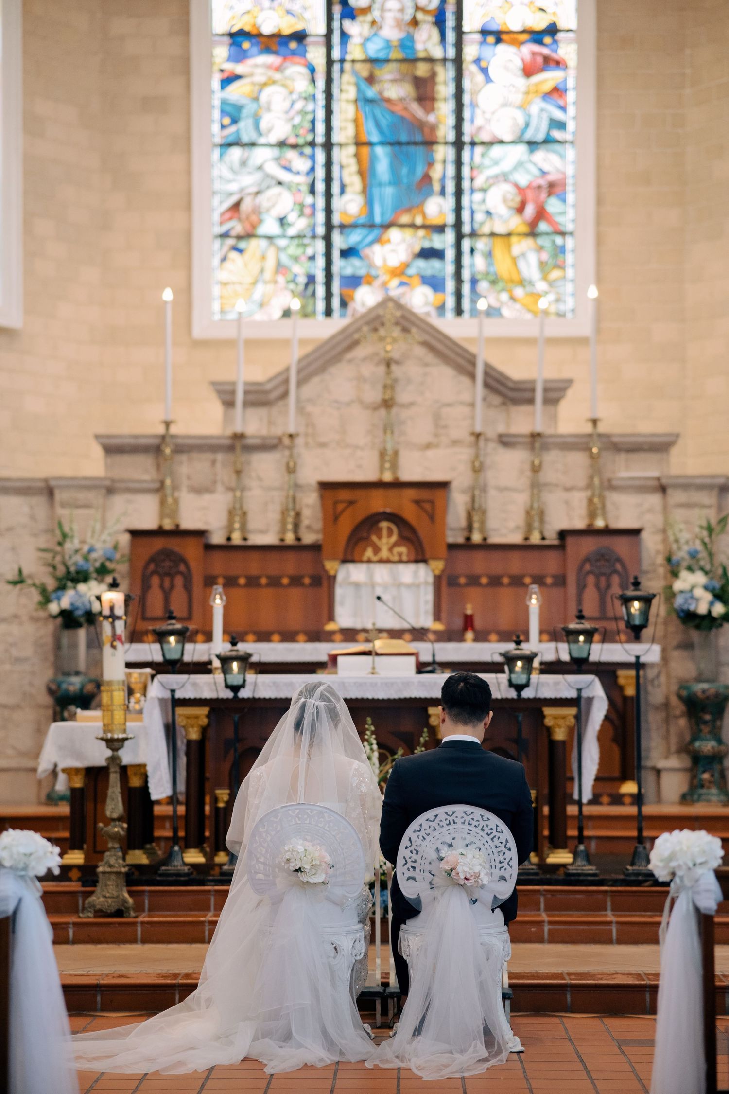 View from the aisle of a bride and groom kneeling at the altar, framed by white floral pew decorations