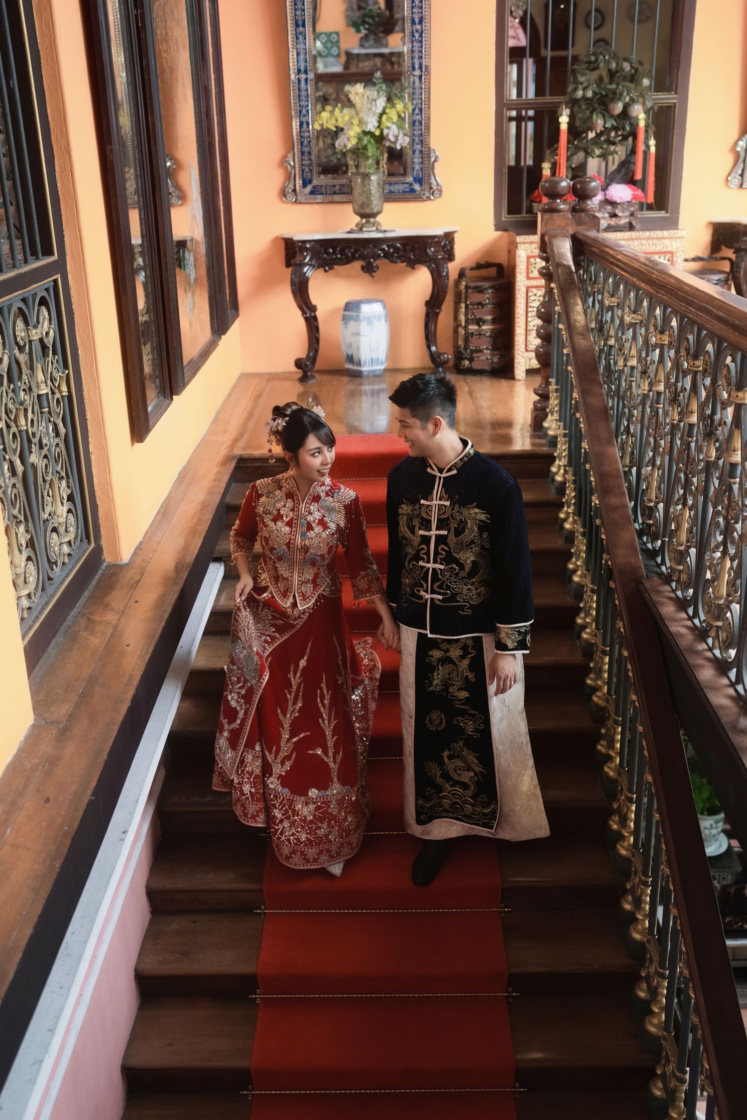 Couple in traditional Chinese wedding attire pose in a hallway with ornate railings and orange walls.