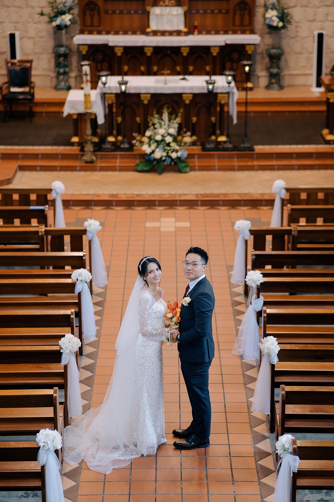 High-angle portrait of a bride and groom standing between pews in a Catholic church