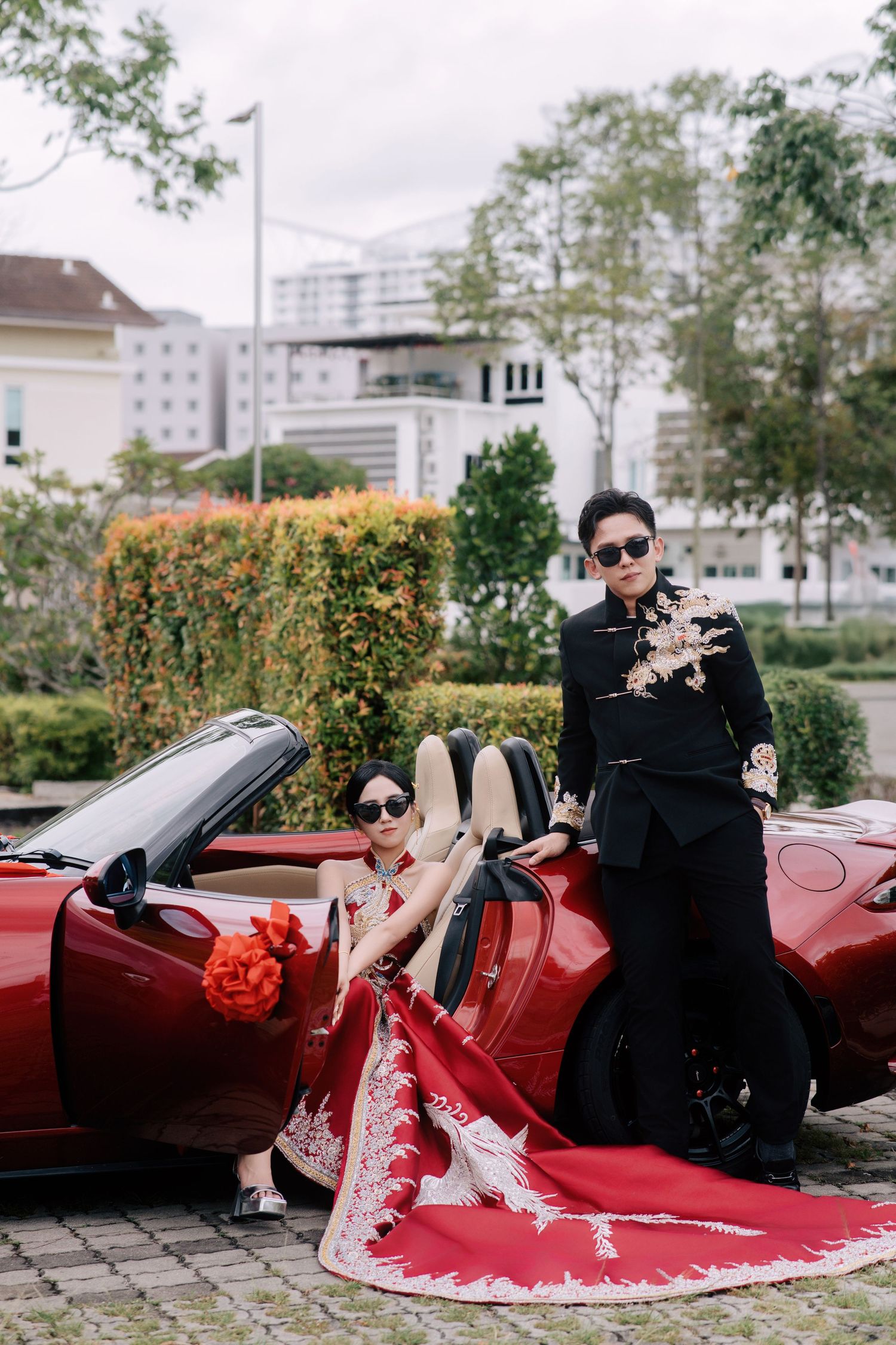 Bride in a red oriental gown sitting by a sports car with her long dress train extended