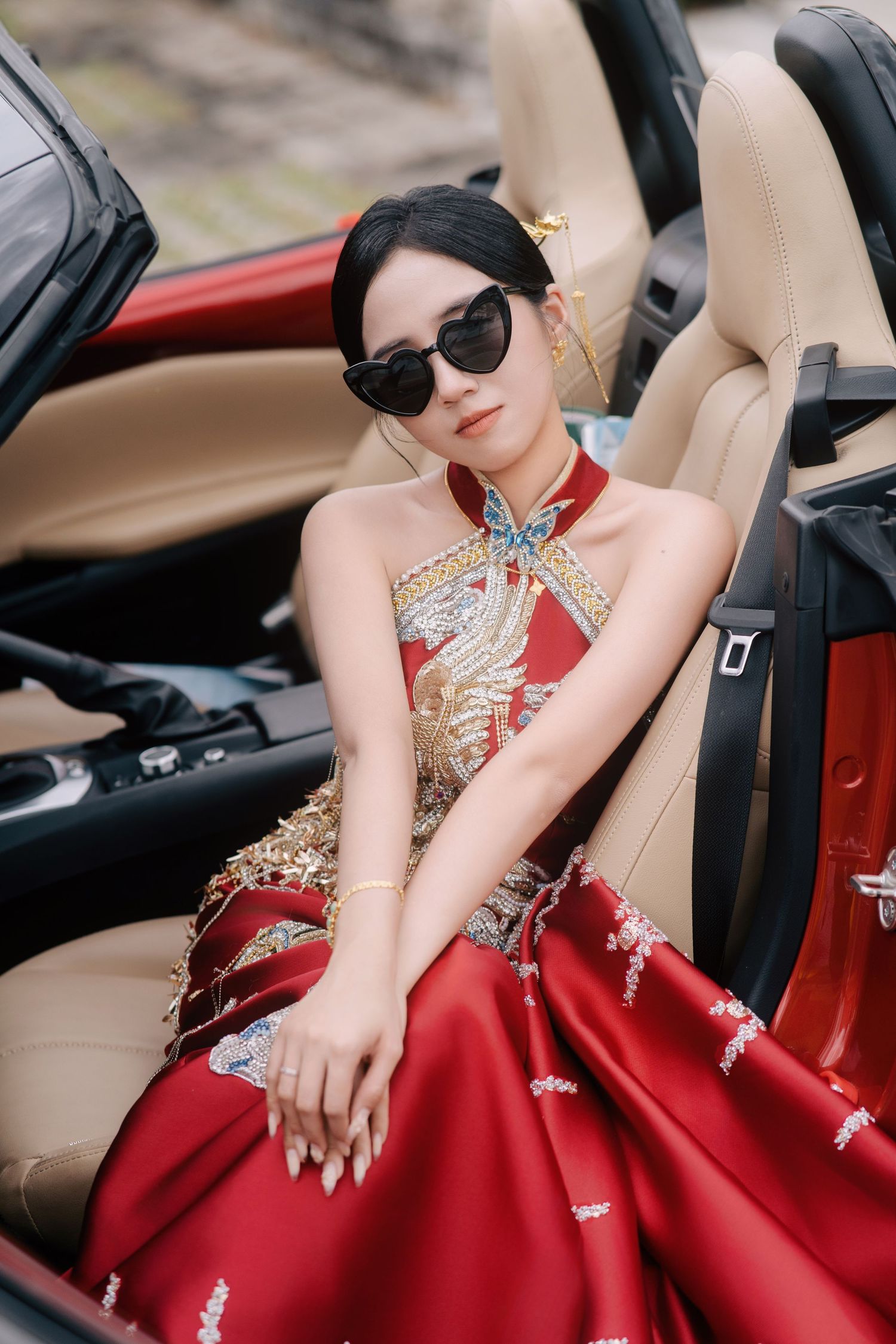Bride wearing heart-shaped sunglasses and a red embroidered dress sitting in a tan car seat