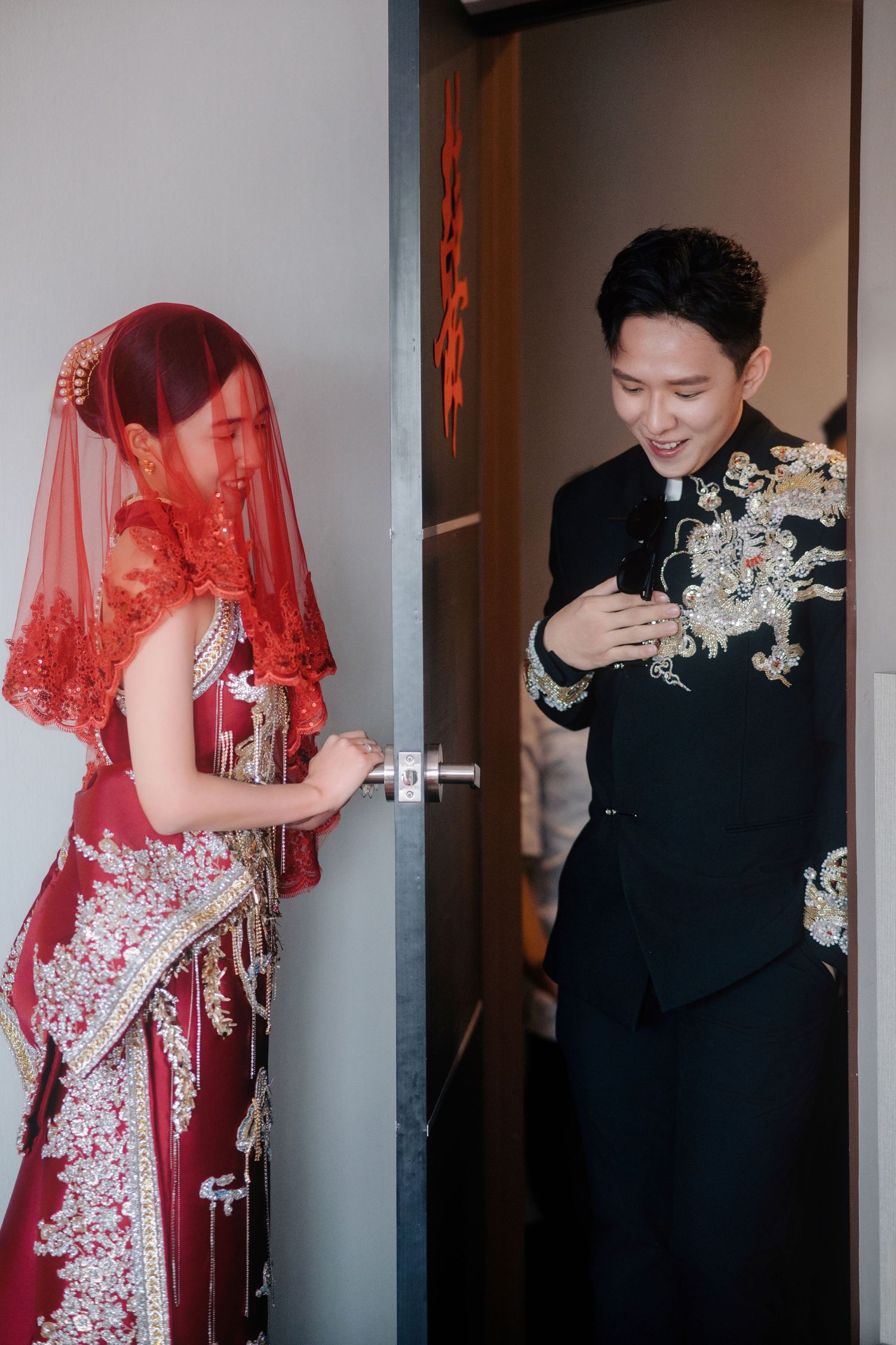 A groom and a veiled bride sharing a smile at the door during a traditional Chinese wedding ceremony