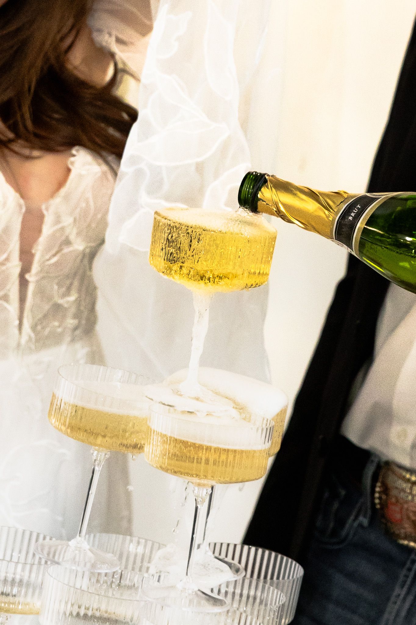 Champagne tower being filled at a wedding reception with bride in white dress in background.