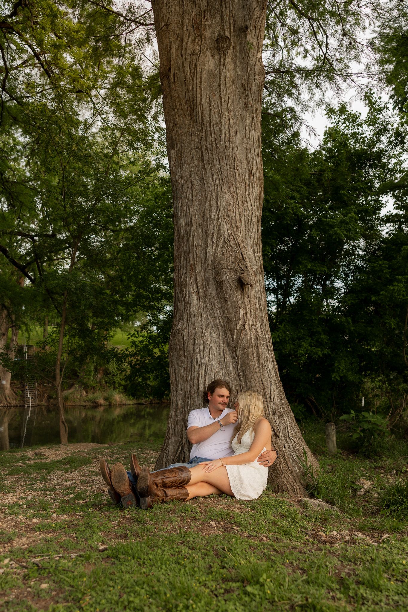 Couple sitting together against a large tree trunk in a lush green park, sharing a romantic outdoor moment.