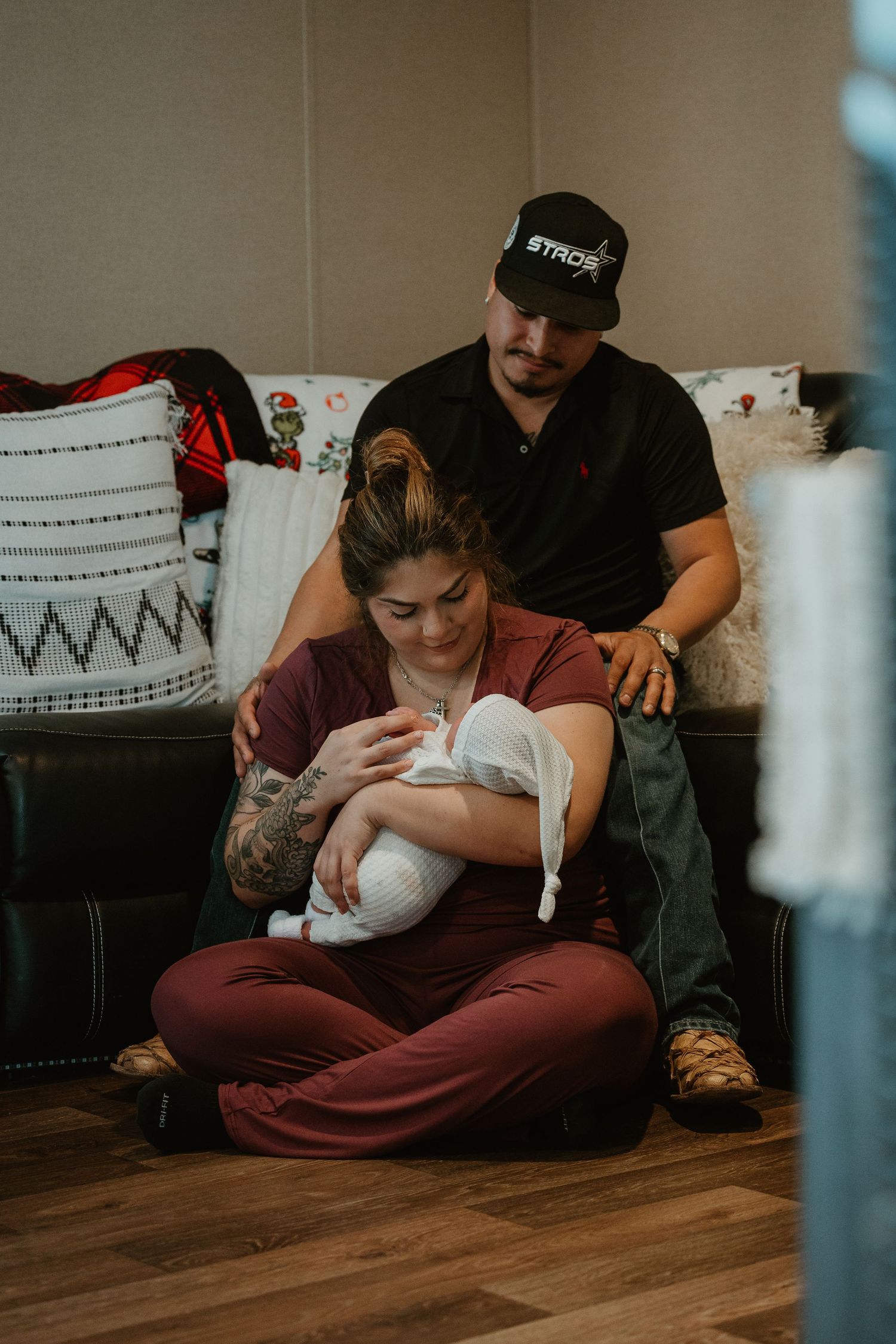 New parents sit on floor cradling newborn baby, mother in maroon outfit with father in black cap looking down lovingly.