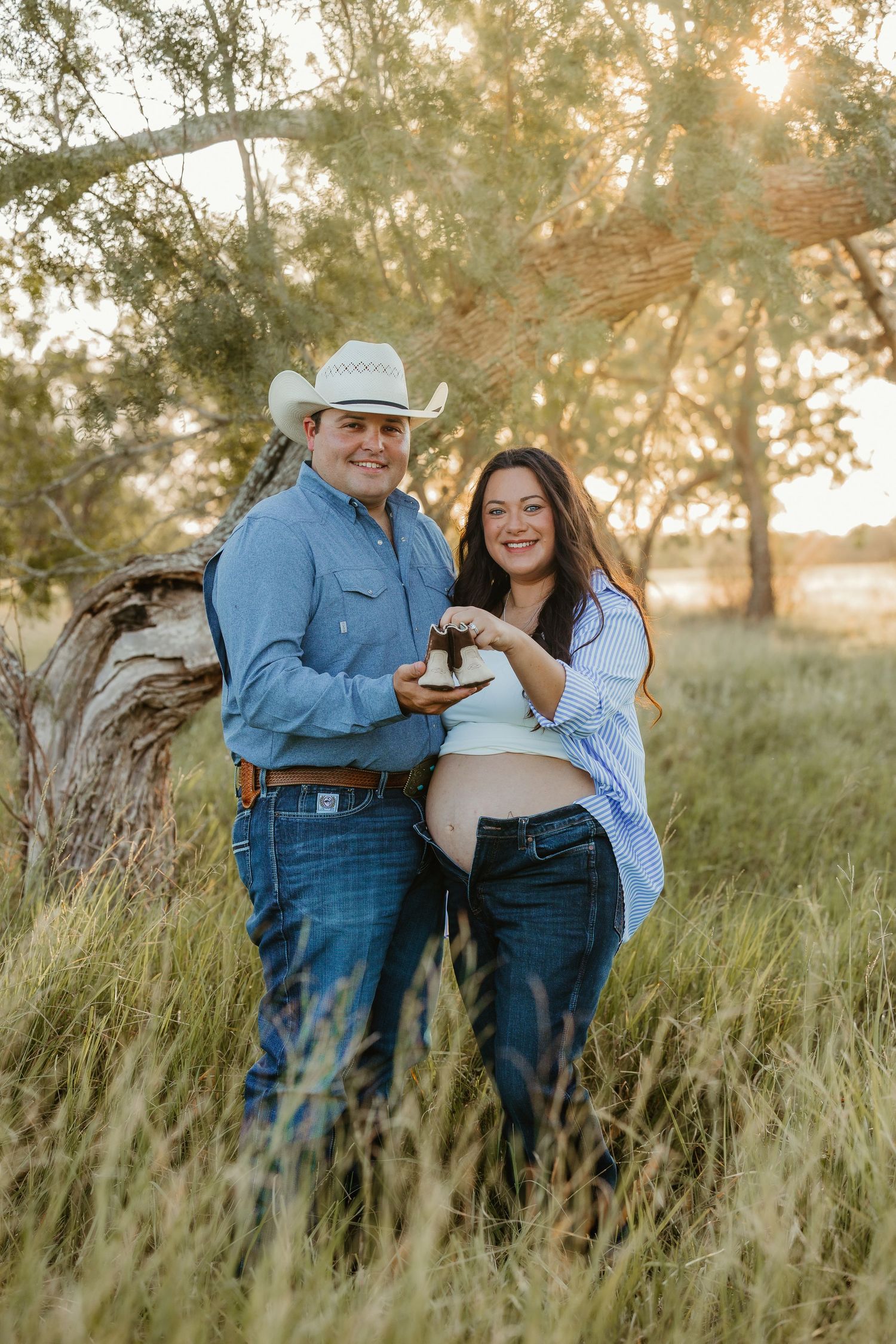 A couple shares a tender moment in tall grass at sunset while wearing casual western attire.