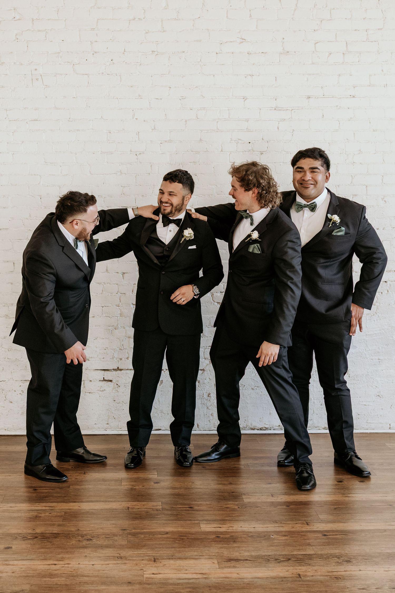 Groomsmen in black suits laughing and posing against a white brick wall.