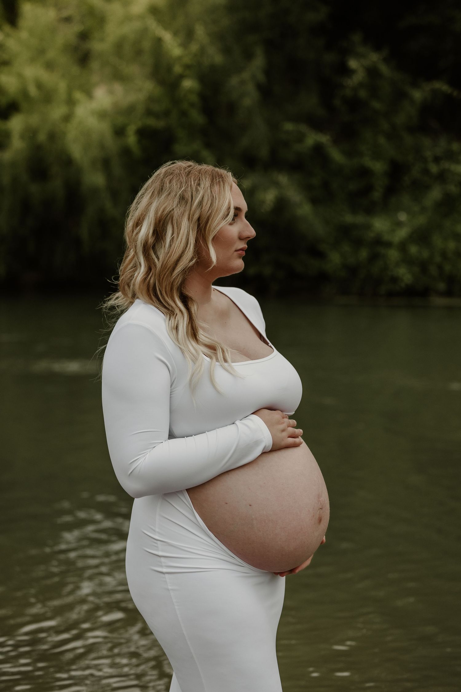 A maternity photo session by a lake captures an expectant mother in a white dress.