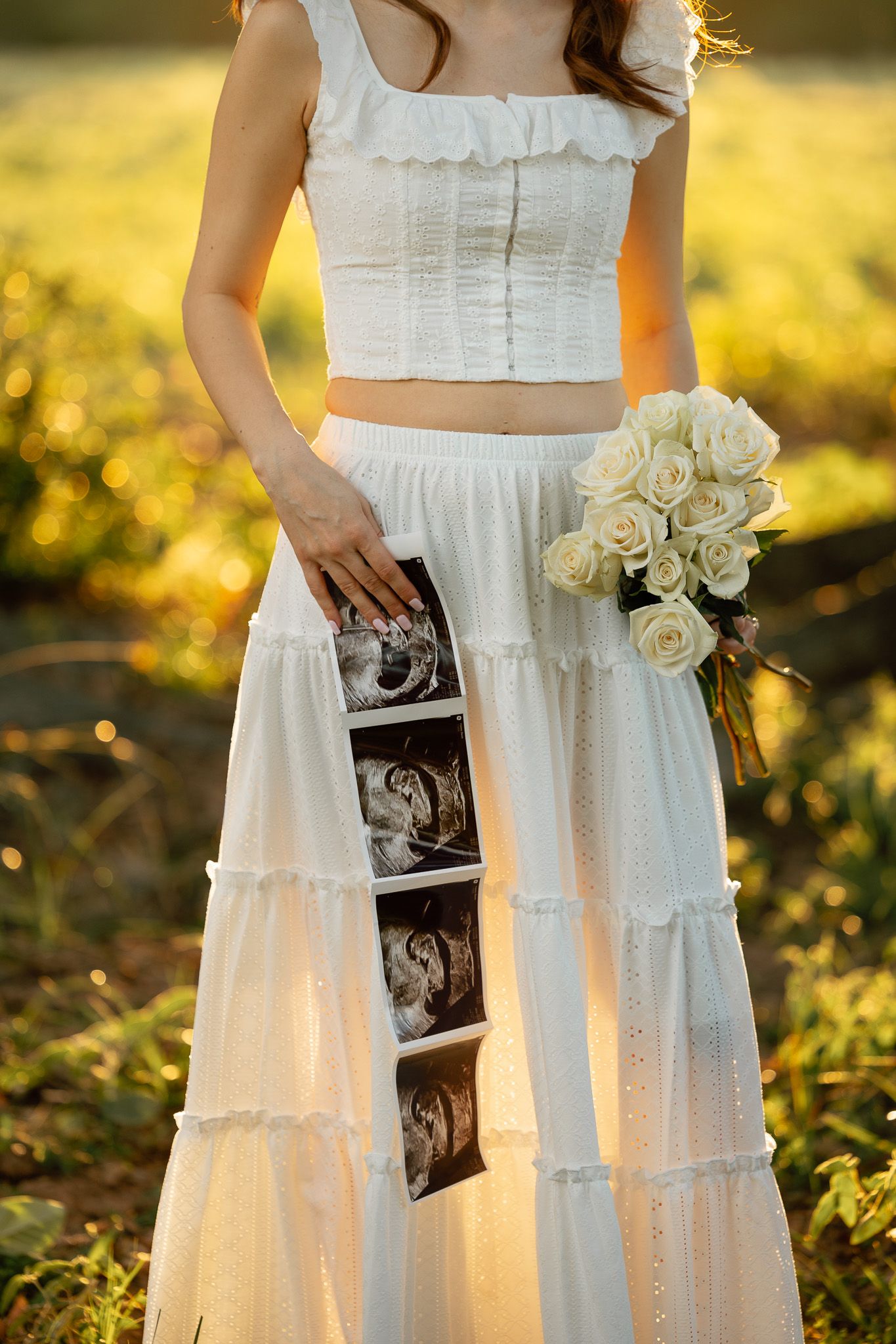 Pregnant woman in white crop top and maxi skirt holding ultrasound photos and white rose bouquet in golden sunlight.