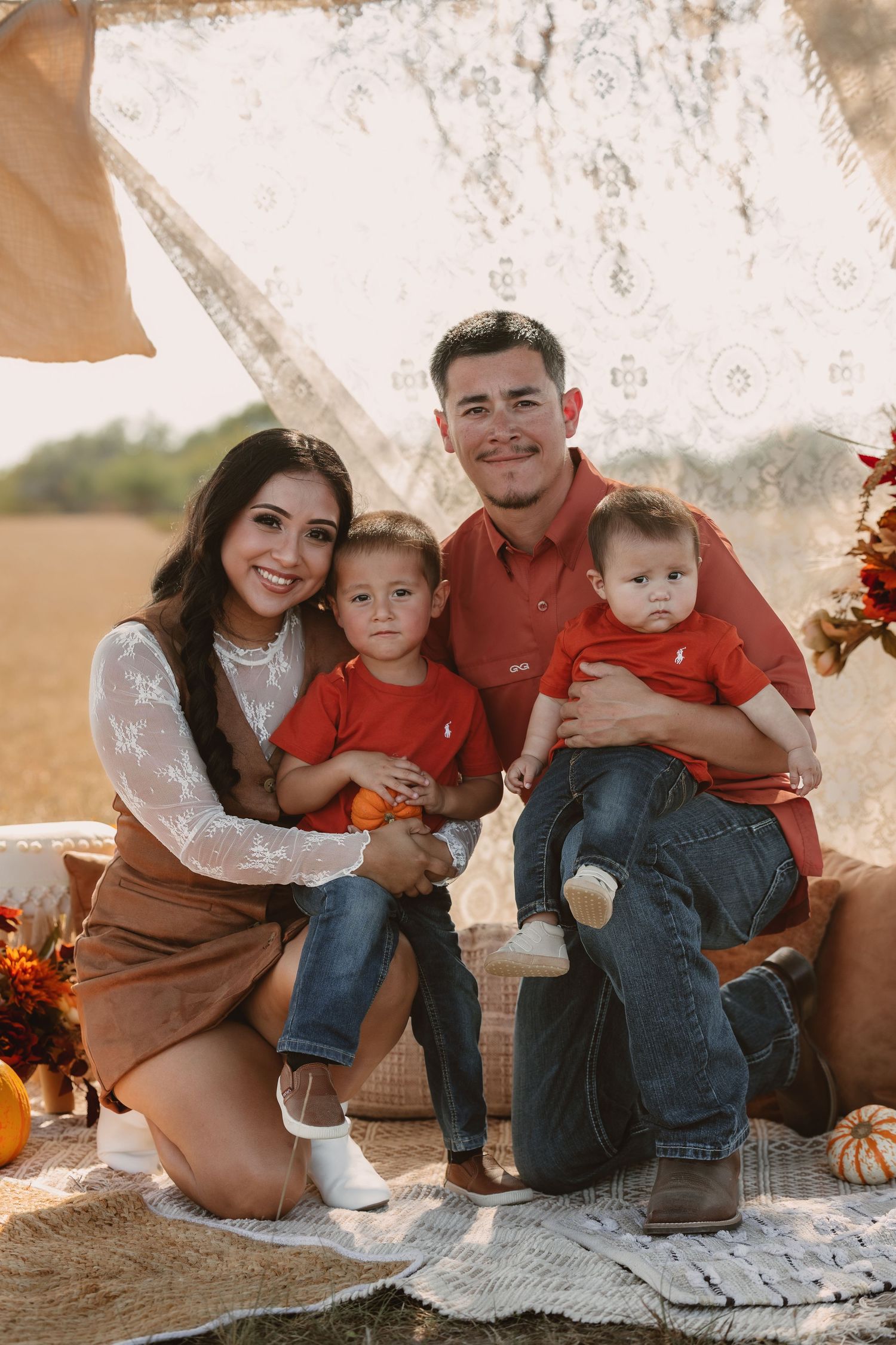 Family of four in matching red outfits posing for a fall-themed outdoor portrait with pumpkins and autumn decor.