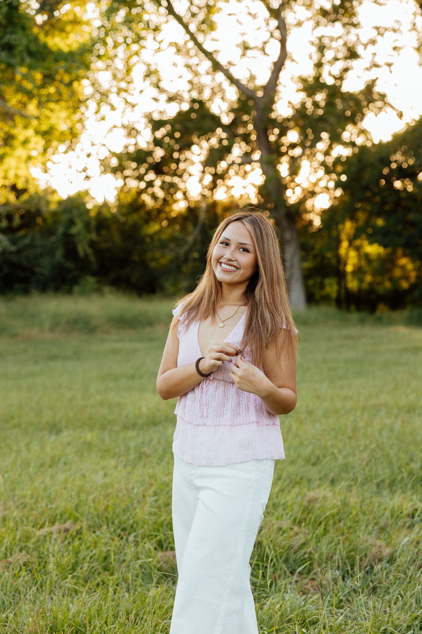Young woman in pink top and white pants smiling outdoors at golden hour with trees in background.