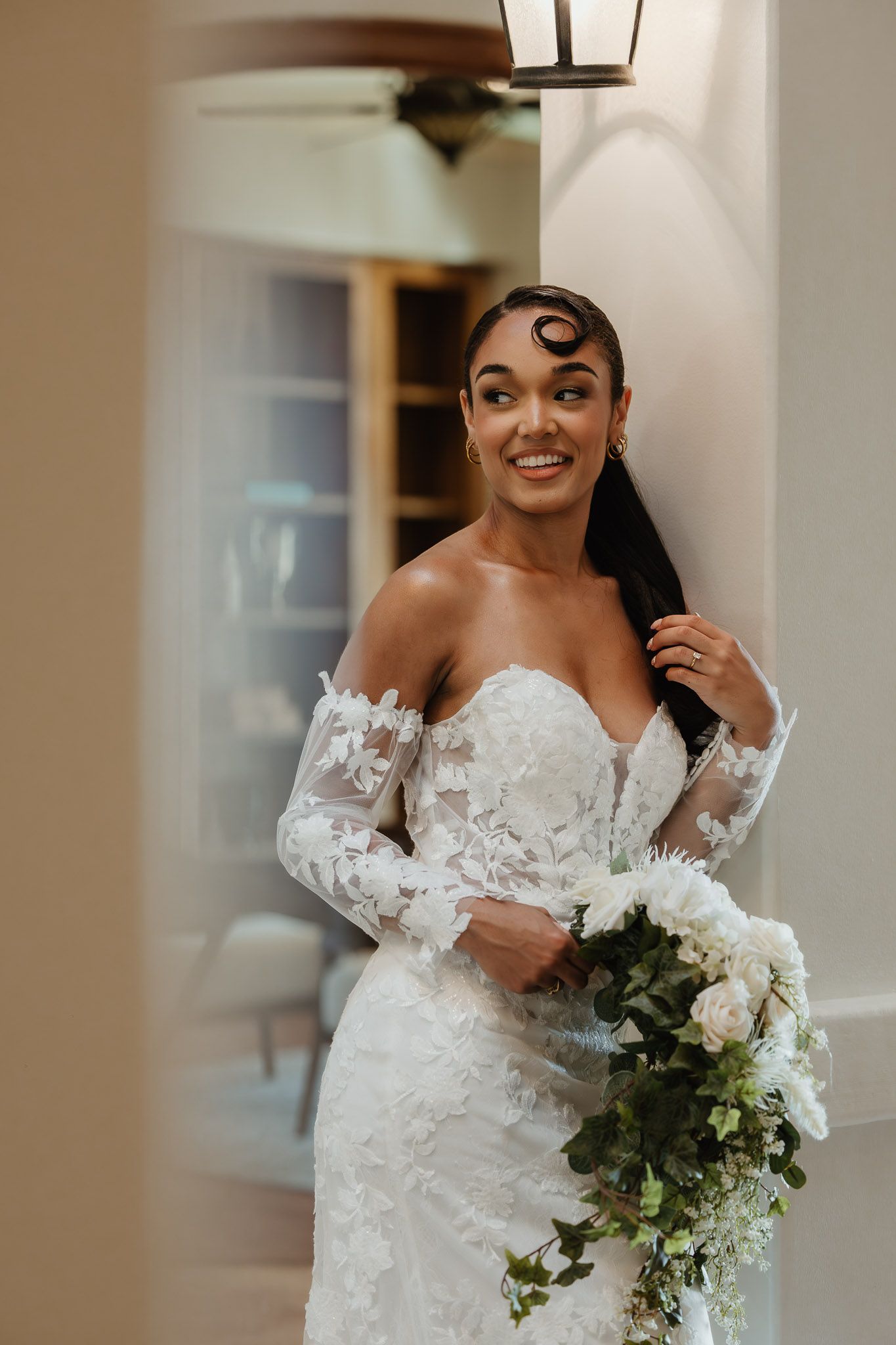 A bride in an elegant off-shoulder lace wedding gown holds a cascading white and green bouquet in an indoor setting.