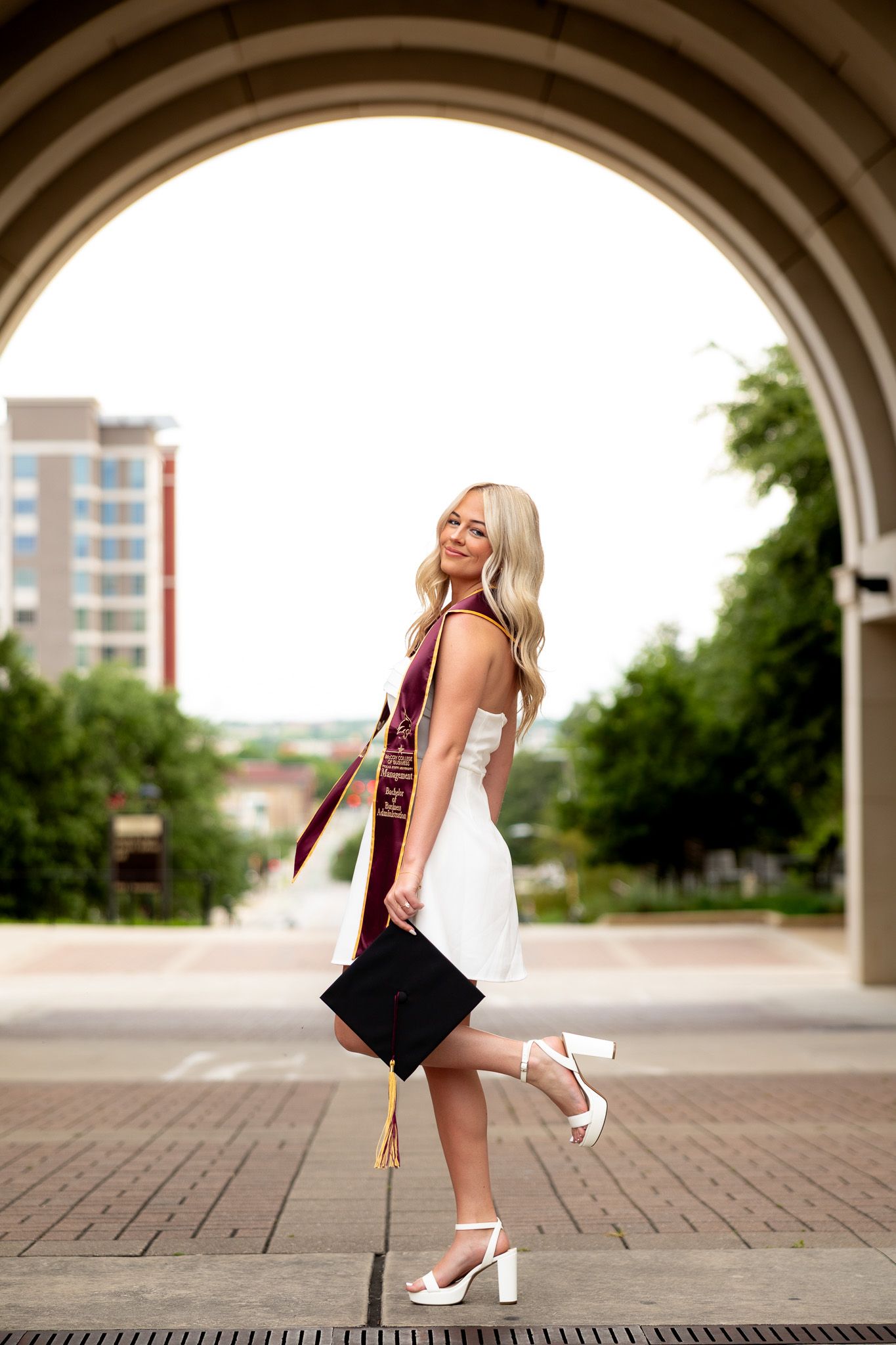 Smiling blonde graduate in white dress holding cap under stone arch, wearing maroon sash and white heels.