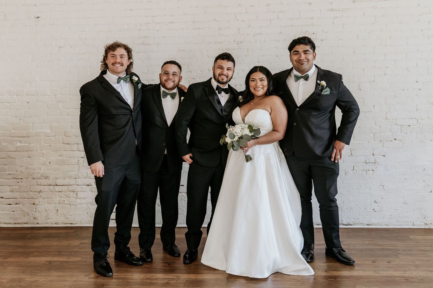 Wedding party poses playfully on hardwood floor against white brick wall in formal attire.