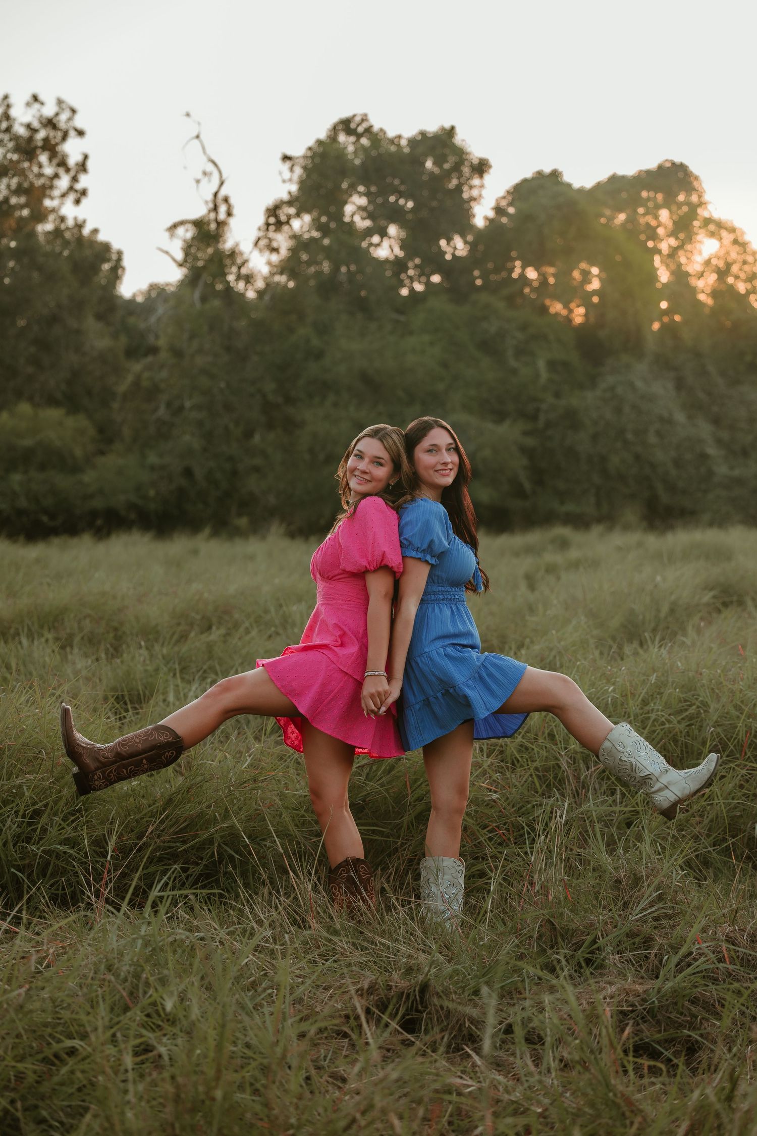 Playful poses in pink and blue dresses amid wild grass during golden hour.