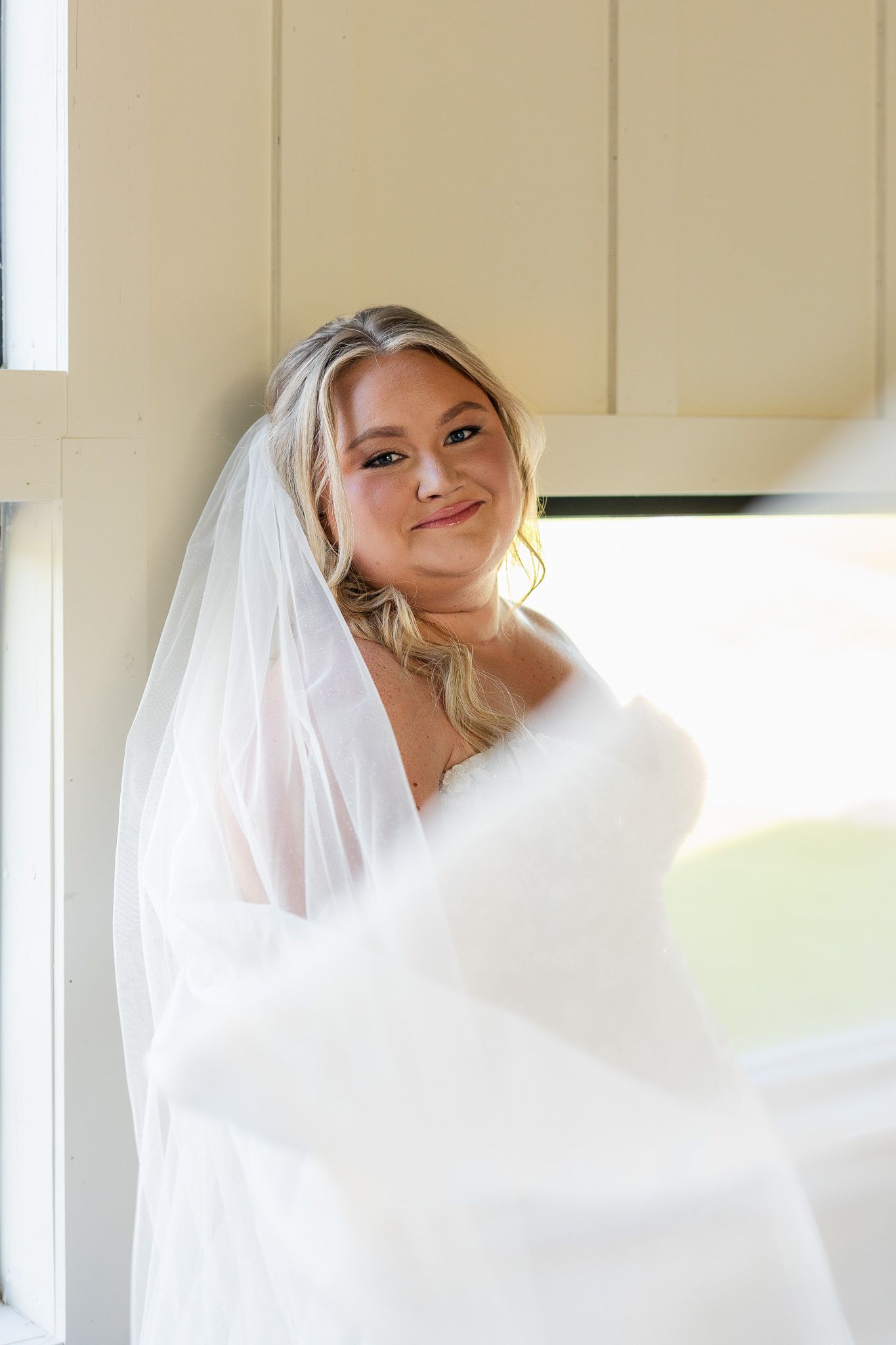 Smiling bride in a white wedding gown and veil posing gracefully by a window on her wedding day.