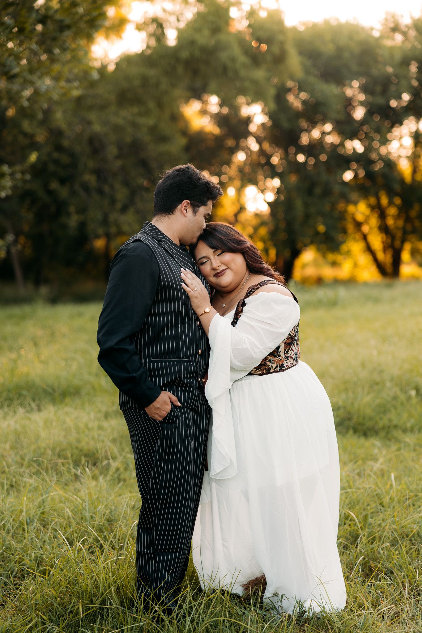Couple embracing in golden hour sunlight in a green field, woman in white dress, man in pinstripe suit.