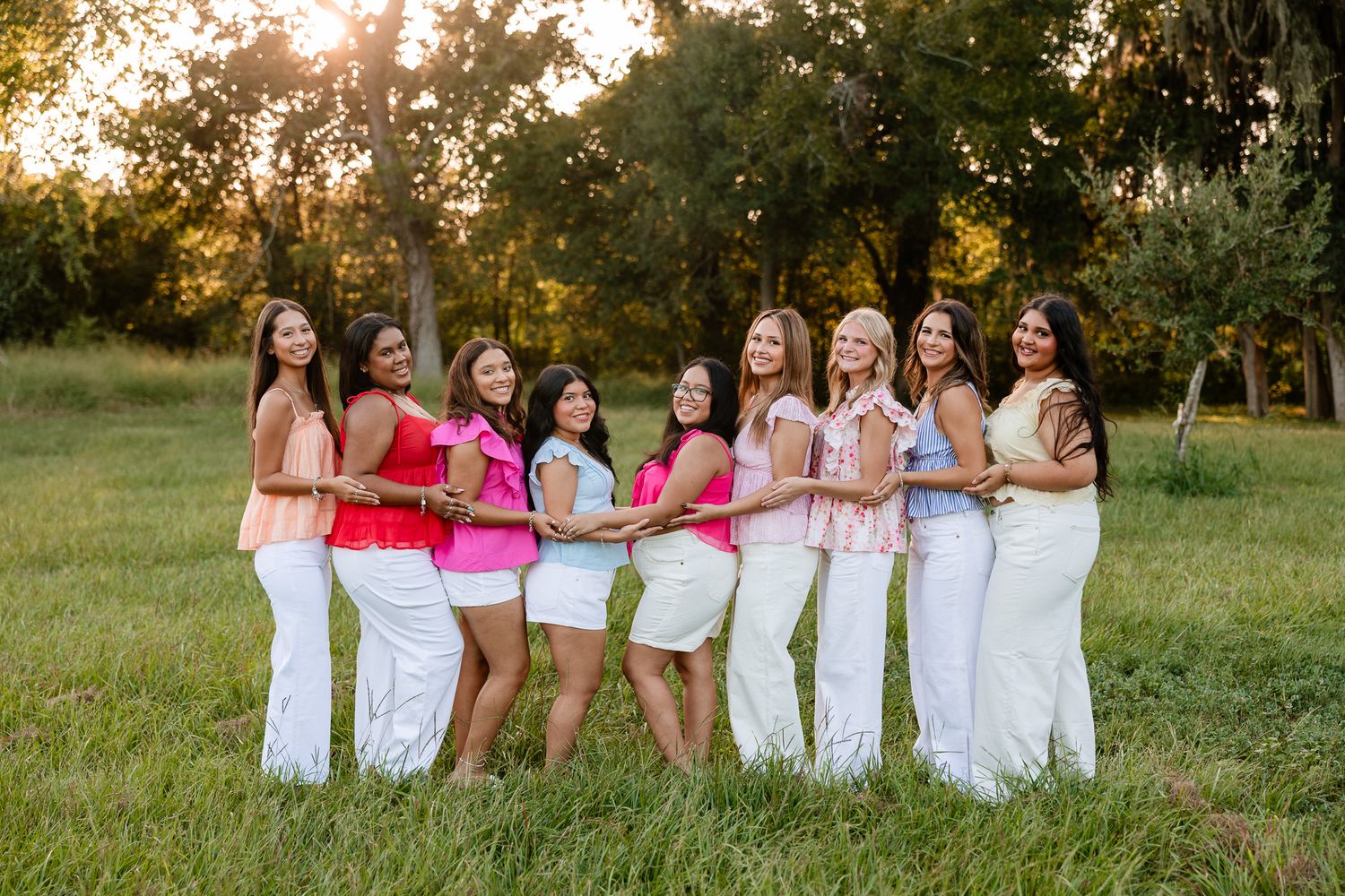 Eight young women in pink tops and white pants pose together in a sunny outdoor field with trees in the background.