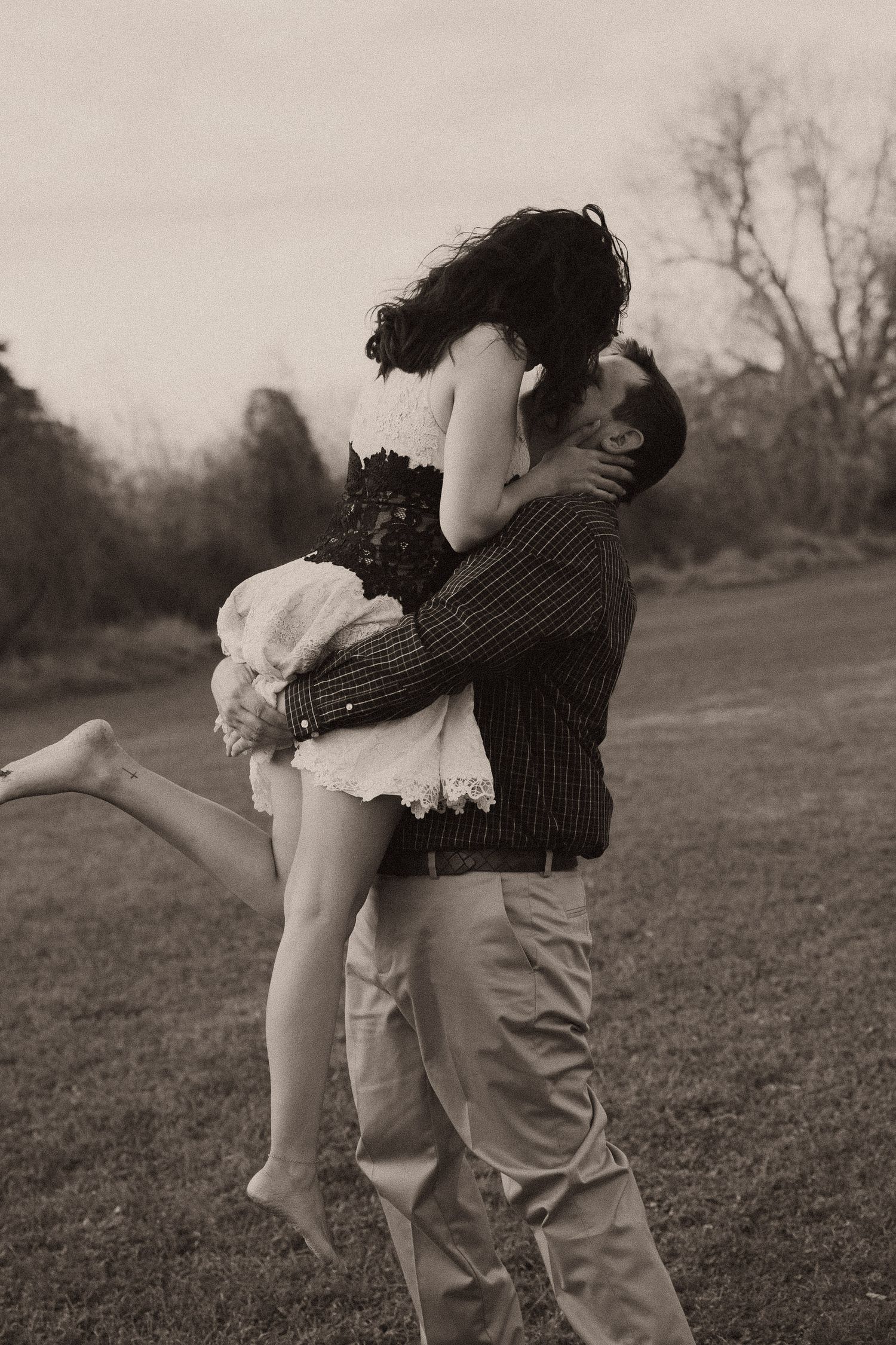A man lifts and kisses a woman in a romantic black and white outdoor photo shoot on a grassy lawn.