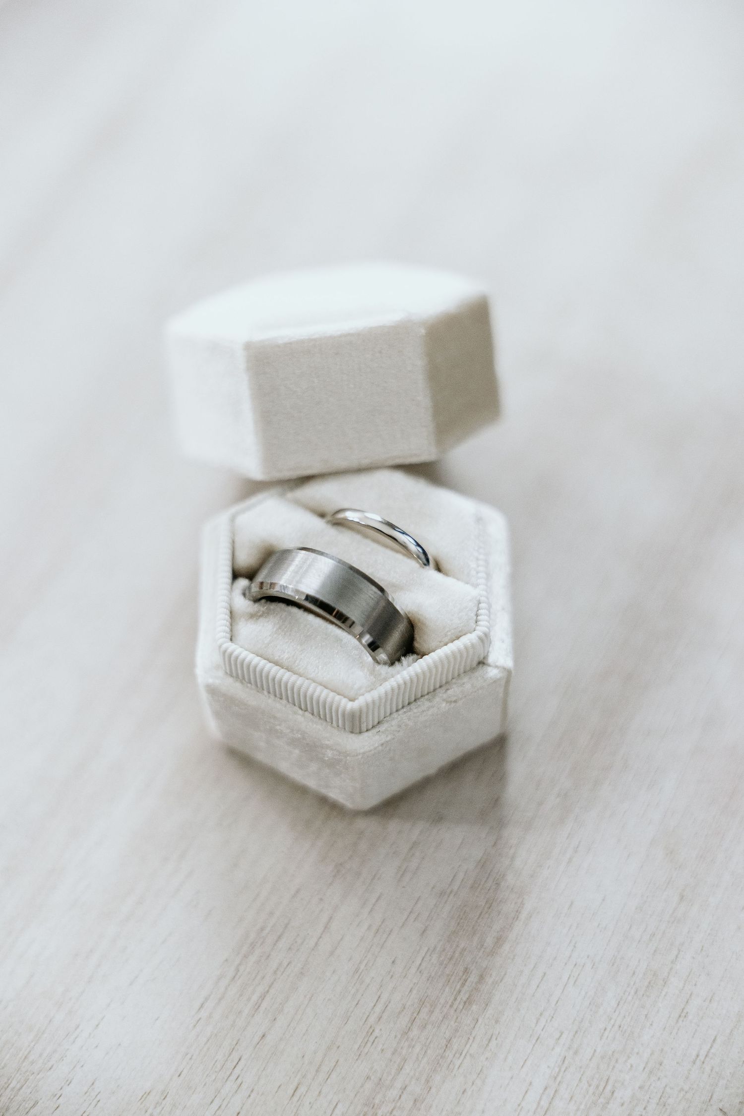 Four minimalist white hexagonal ring boxes with silver wedding bands shown in a row on a light gray wooden surface.